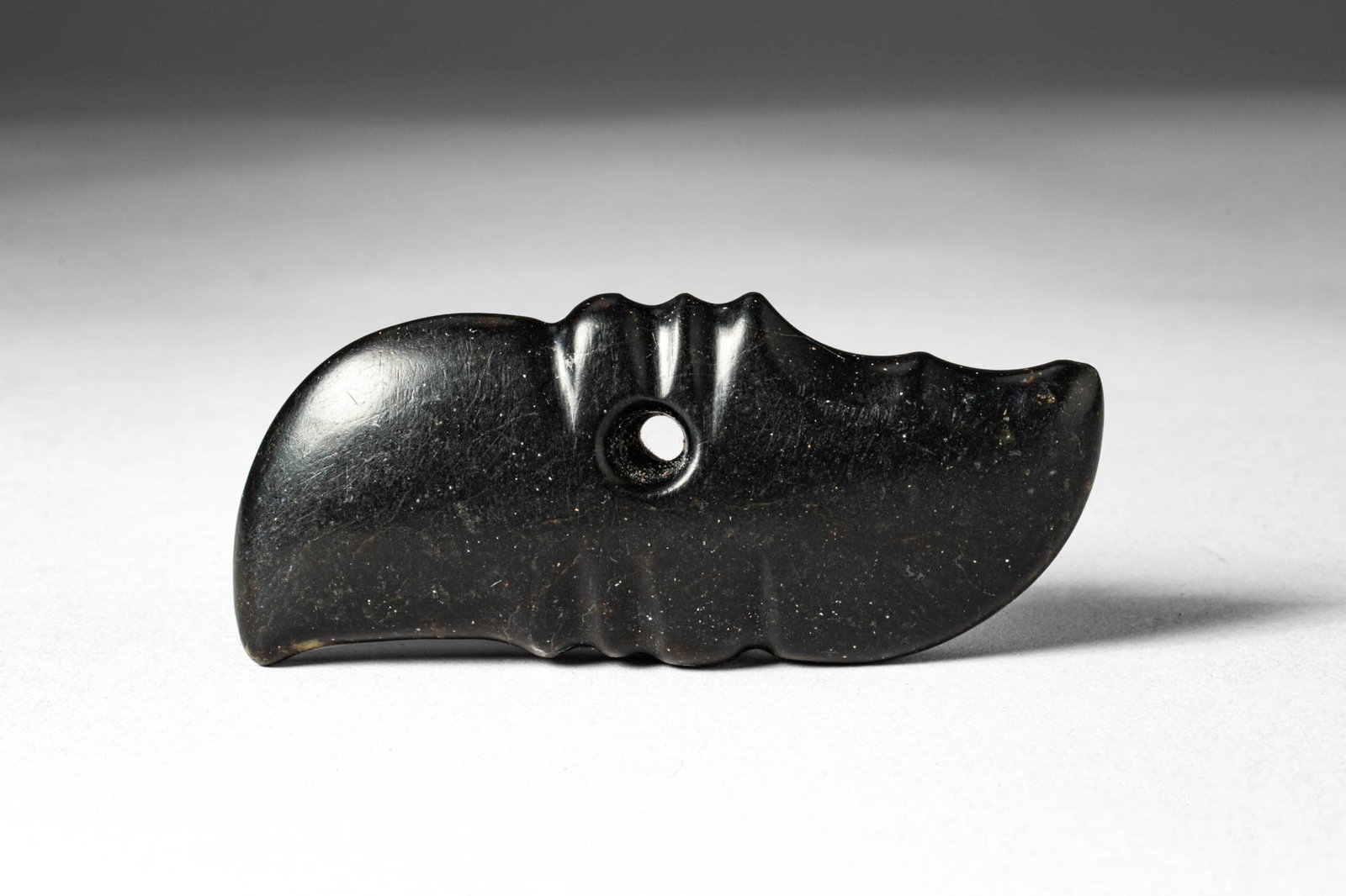 CHINESE JADE PENDANT: . >A pendant carved in dark jade of elongated, slightly curved form, the surface modelled with shallow ridged bands and pierced with a circular aperture for suspension. The stone is finely polished, d