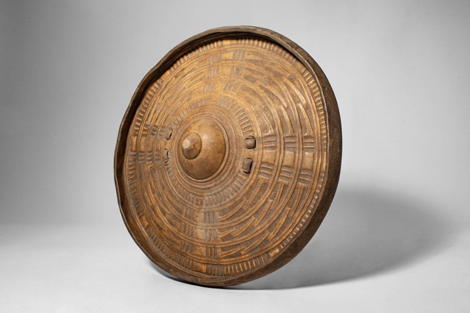 SOMALIAN WOODEN SHIELD: Ca. 19th century AD. > A shield of circular form, the convex wooden body centred by a raised boss and decorated overall with finely incised concentric bands and geometric motifs. The carved ornament i