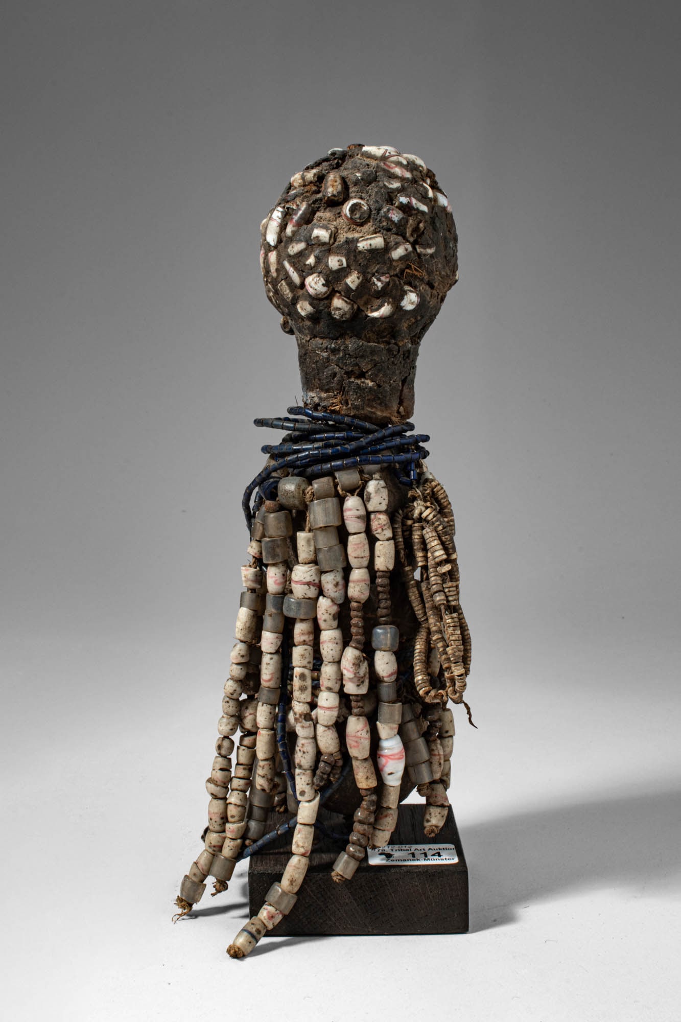 AFRICAN FERTILITY DOLL: Ca. late 19th - early 20th century AD. >A fertility doll of wood and organic materials, the figure with a rounded head encrusted with shells and small applied elements, above a cylindrical neck encirc