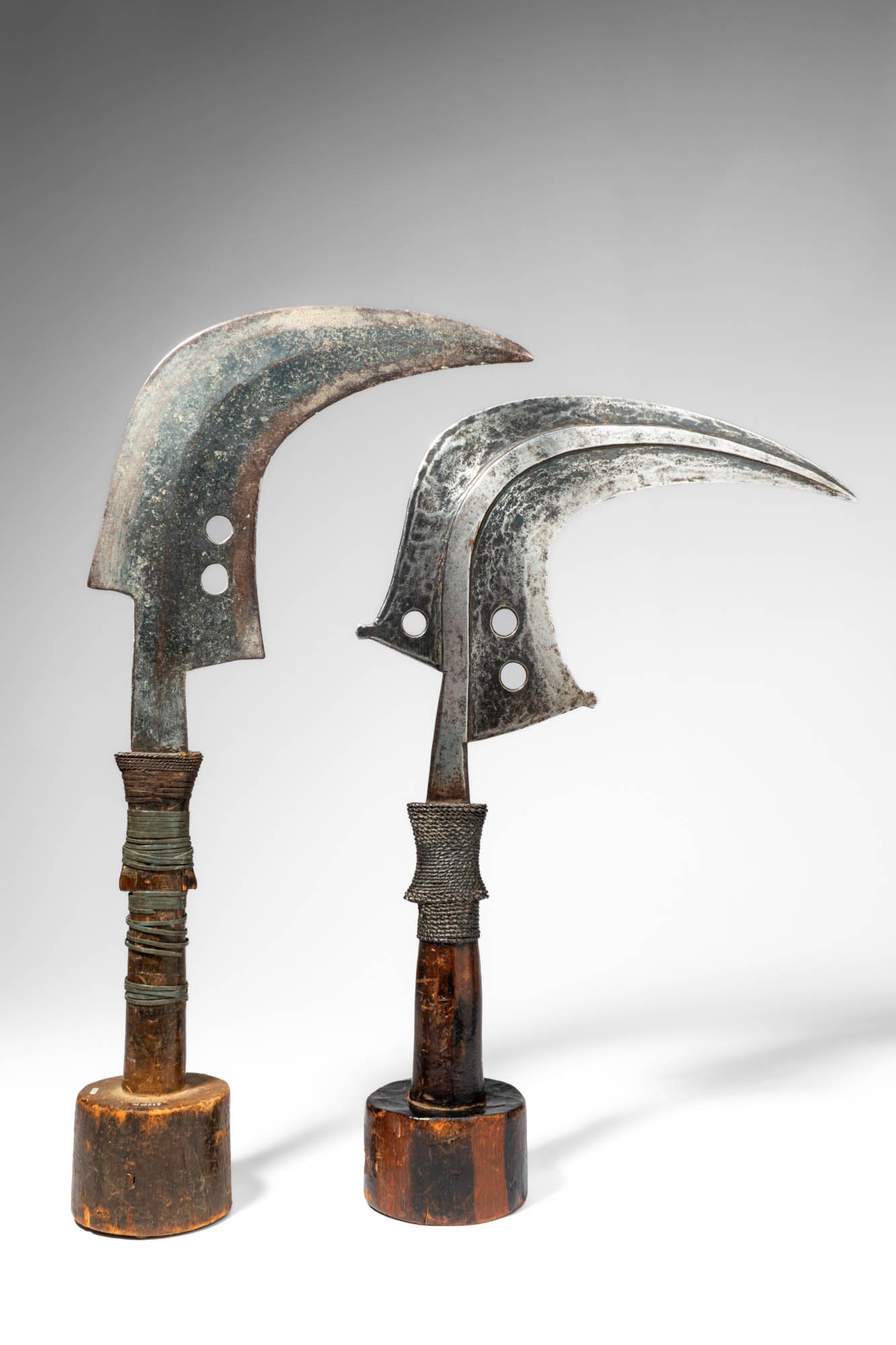 PAIR OF MANGBETU TRUMBASH SICKLE KNIVES: Ca. 20th century AD. >A pair of Mangbetu trumbash knives, each with a distinctive crescent-shaped iron blade mounted on a short handle. The blades are of pronounced curved form, tapering to sharp poin