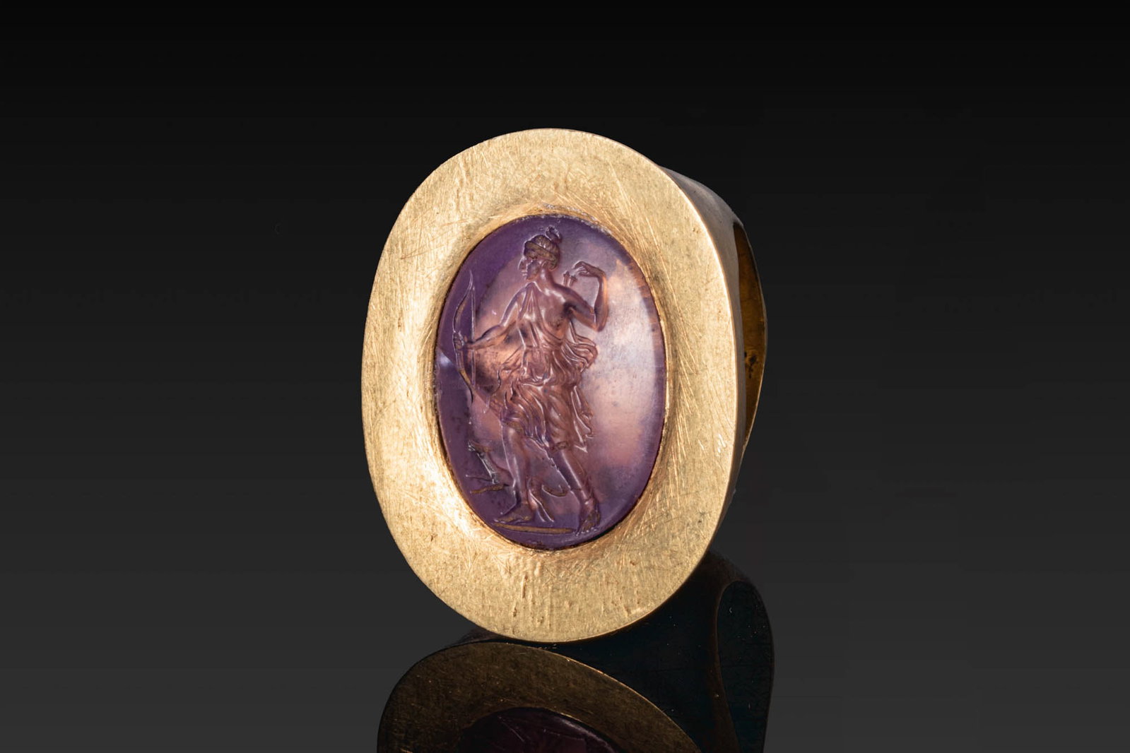 STUNNING HELLENISTIC GOLD RING WITH AMETHYST INTAGLIO OF ARTEMIS - HUGE: Ca. 200 - 1 BC. A gold ring with a broad, rounded hoop and an oval bezel plate set with an amethyst intaglio of Artemis, standing in profile on a baseline, with a quiver over her shoulder as she reach