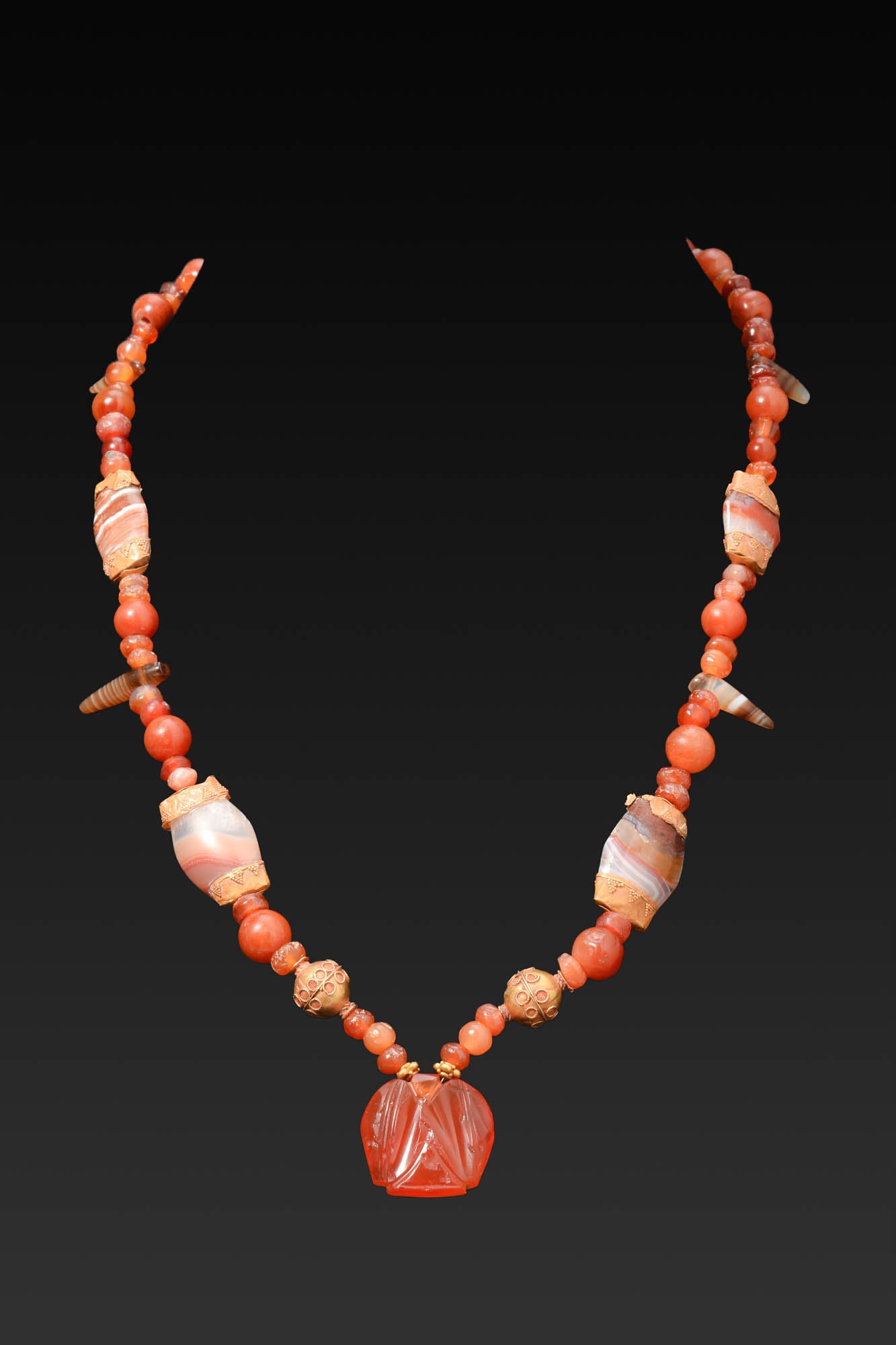 GRAECO-BACTRIAN GOLD, CARNELIAN AND AGATE NECKLACE: Ca. 300 BC. A necklace with a strand of carnelian beads, interspersed with banded agate barrel-shaped spacers with gold collars, gold spherical beads, and tapering agate drops, centred by a carved aga