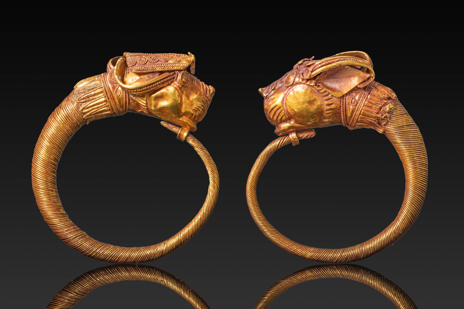 MATCHED PAIR OF GREEK GOLD EARRINGS WITH ZOOMORPHIC FINIALS: Ca. 450 - 250 BC. A matched pair of gold earrings, each formed as a tapering hoop wrapped with fine wire and terminating in a sculptural zoomorphic head with the horns curving backwards over the crown