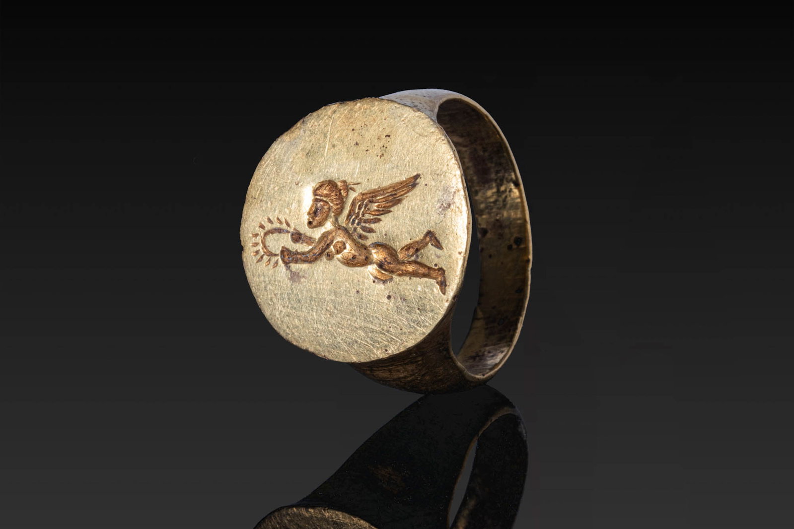 GREEK SOLID GOLD RING WITH FLYING EROS: Ca. 300 BC. A gold ring with a flat-section, tapering hoop and a circular flat bezel engraved with a flying Eros in his left profile, holding a wreath before him. For similar see: The British Museum, 