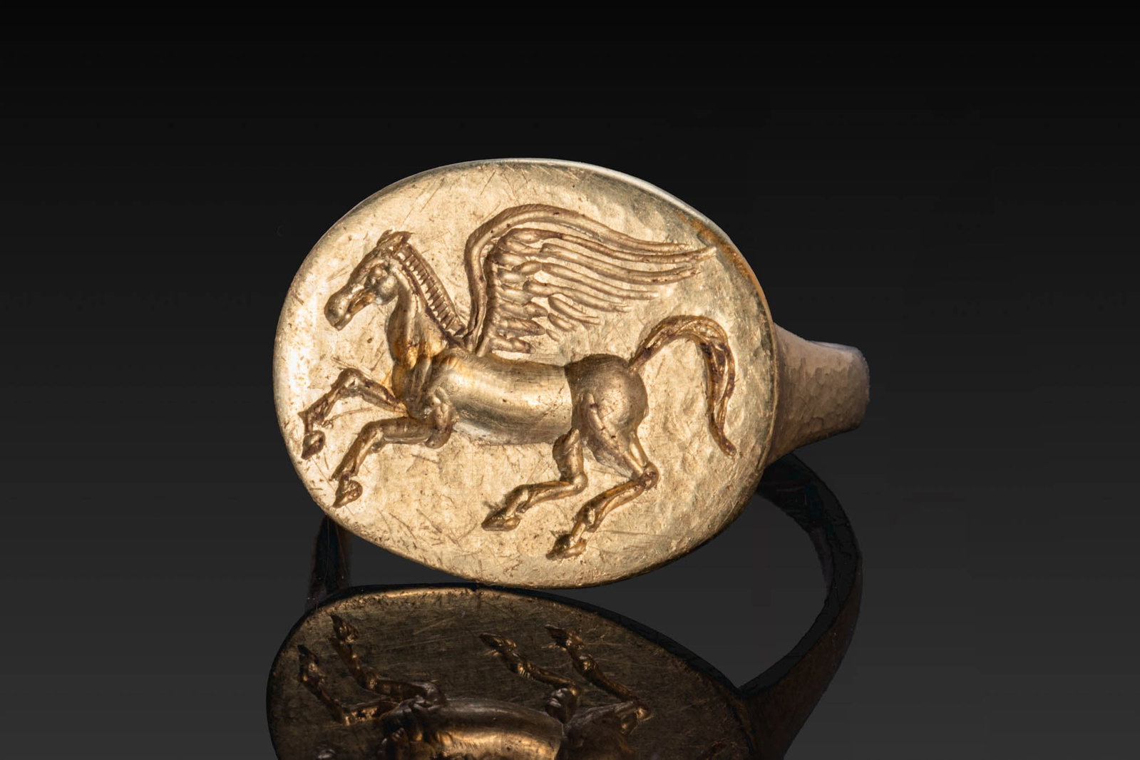 GREEK HELLENISTIC GOLD RING WITH PEGASUS: Ca. 400 BC. A gold ring with a flat-section hoop broadening to a large oval bezel engraved with Pegasus advancing left with forelegs raised, wings outspread, and tail curled upward. Size: D:17.73mm / 