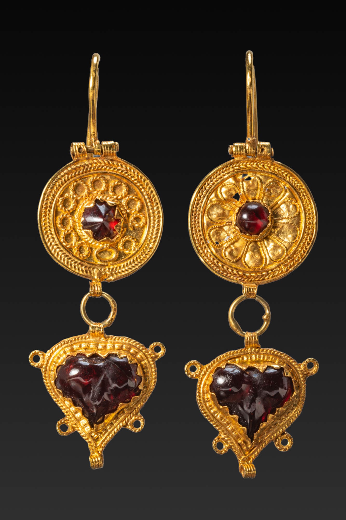 MATCHED PAIR OF HELLENISTIC GOLD EARRINGS WITH GARNETS: Ca. 150 BC. A matched pair of gold earrings, each with a plain hook above a circular medallion framed by braided wire and set at the centre with a garnet. One medallion is decorated with a rosette of 