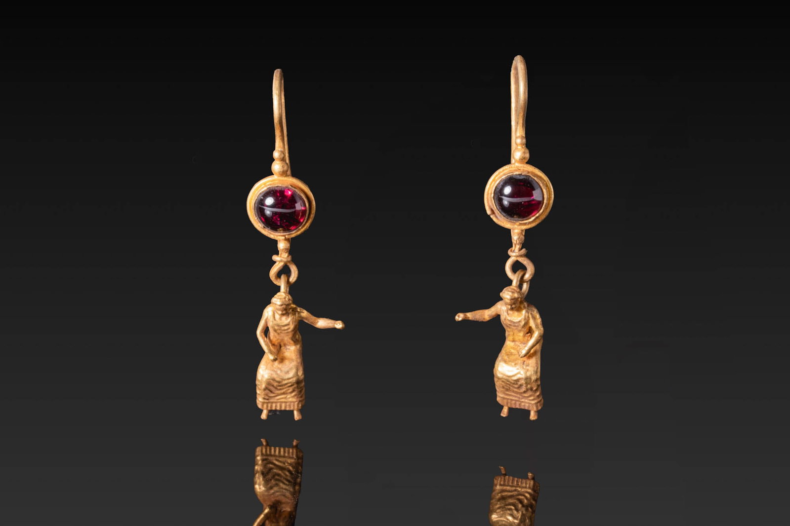 MATCHED PAIR OF HELLENISTIC GOLD EARRINGS WITH GARNET CABOCHONS AND FEMALE FIGURES: Ca. 300 - 100 BC. A matched pair of gold earrings, each with a hook above a circular garnet cabochon in a plain mount, with granules above and below, suspending a seated female figure in a long dress 