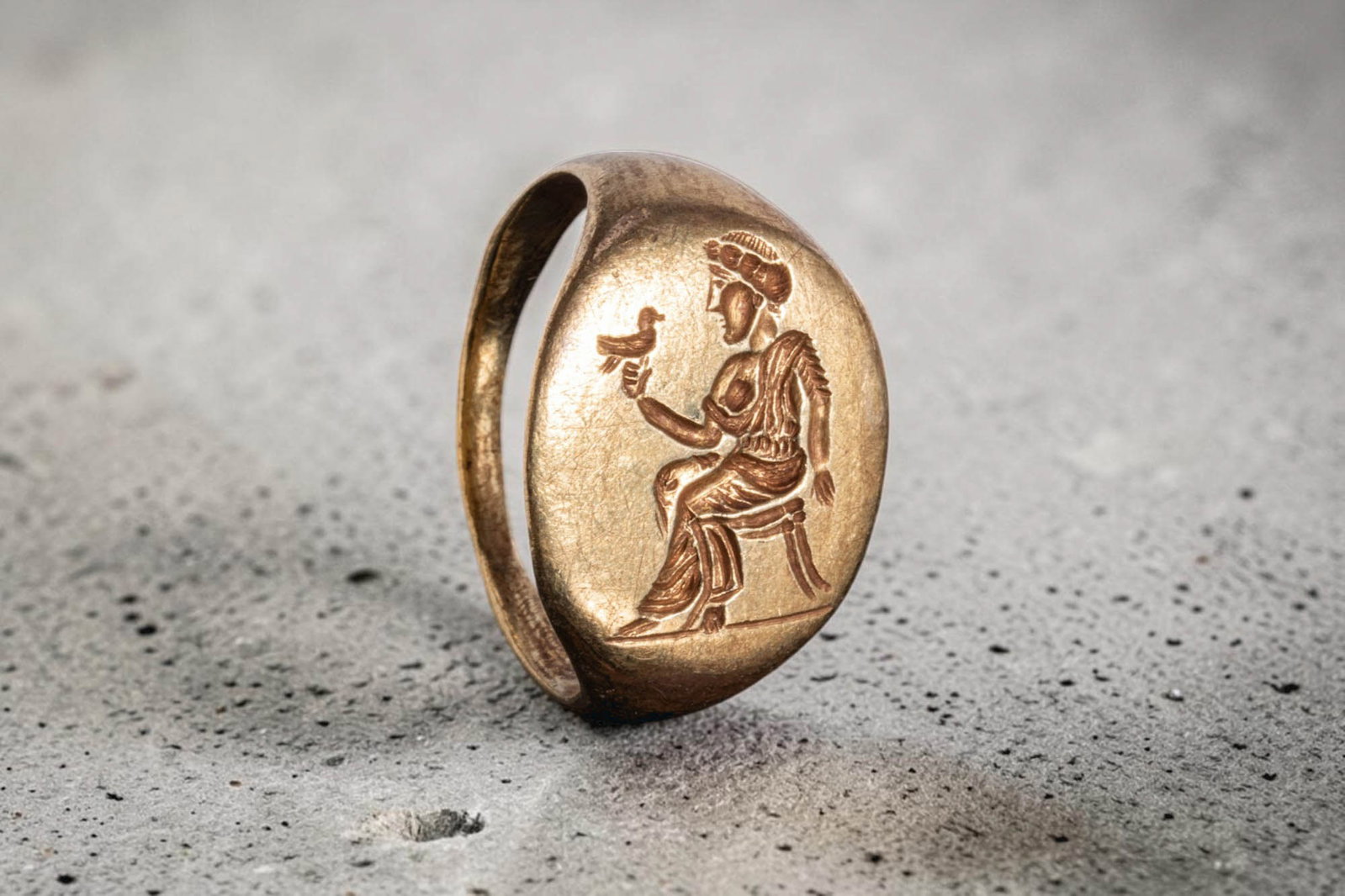 STUNNING HELLENISTIC SOLID GOLD RING WITH SEATED QUEEN HOLDING A DOVE: Ca. 300 - 100 BC. A gold ring with a slender flat-section hoop widening to a large oval bezel. The bezel is engraved with a seated queen in profile on a stool, wearing a long draped garment and a diad