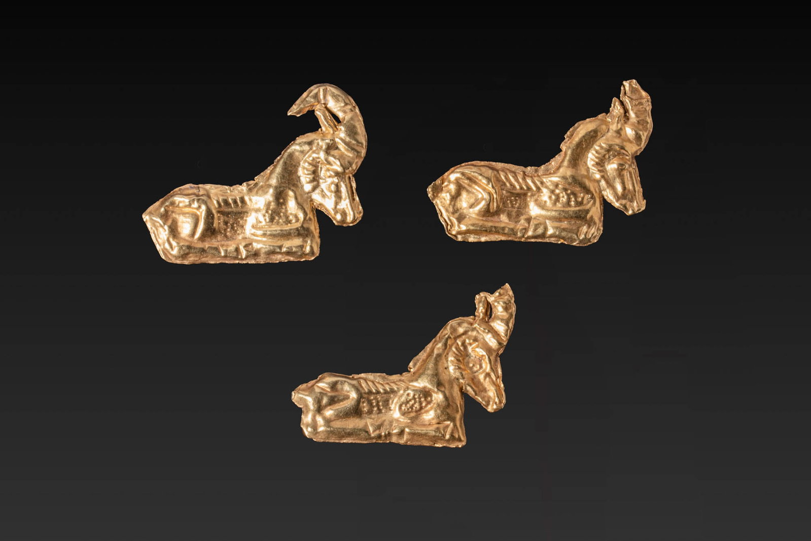 COLLECTION OF GREEK PERIOD HIGH KARAT GOLD IBEX APPLIQUES: Ca. 500 BC. A collection of three gold appliques, each modelled as a recumbent ibex with swept horns and detailing to the head, shoulder, and haunch, with four tubular loops to the reverse for attachm