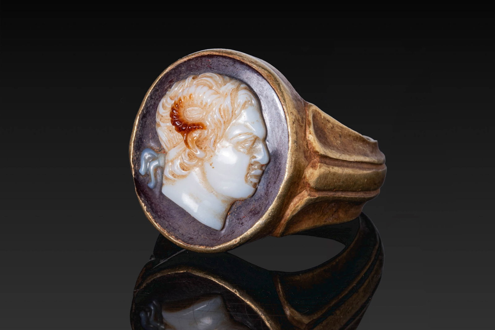 HELLENISTIC AGATE CAMEO OF A RULER AS ZEUS AMMON IN GOLD RING: Ca. 300 - 100 BC. An agate cameo carved with the profile bust of a ruler depicted as Zeus Ammon, with a ram’s horn at the temple and a fillet tied at the back of the curly hair, set in a possibly la