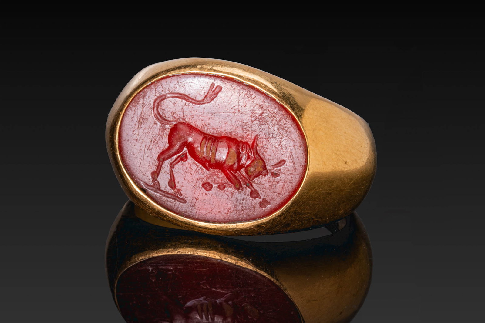 HELLENISTIC GOLD RING WITH CARNELIAN INTAGLIO OF A BULL: Ca. 100 BC. A gold ring with a broad hoop expanding to an oval bezel set with a carnelian intaglio engraved with a bull lowering its head and forelegs, with a curved tail above and a groundline below.