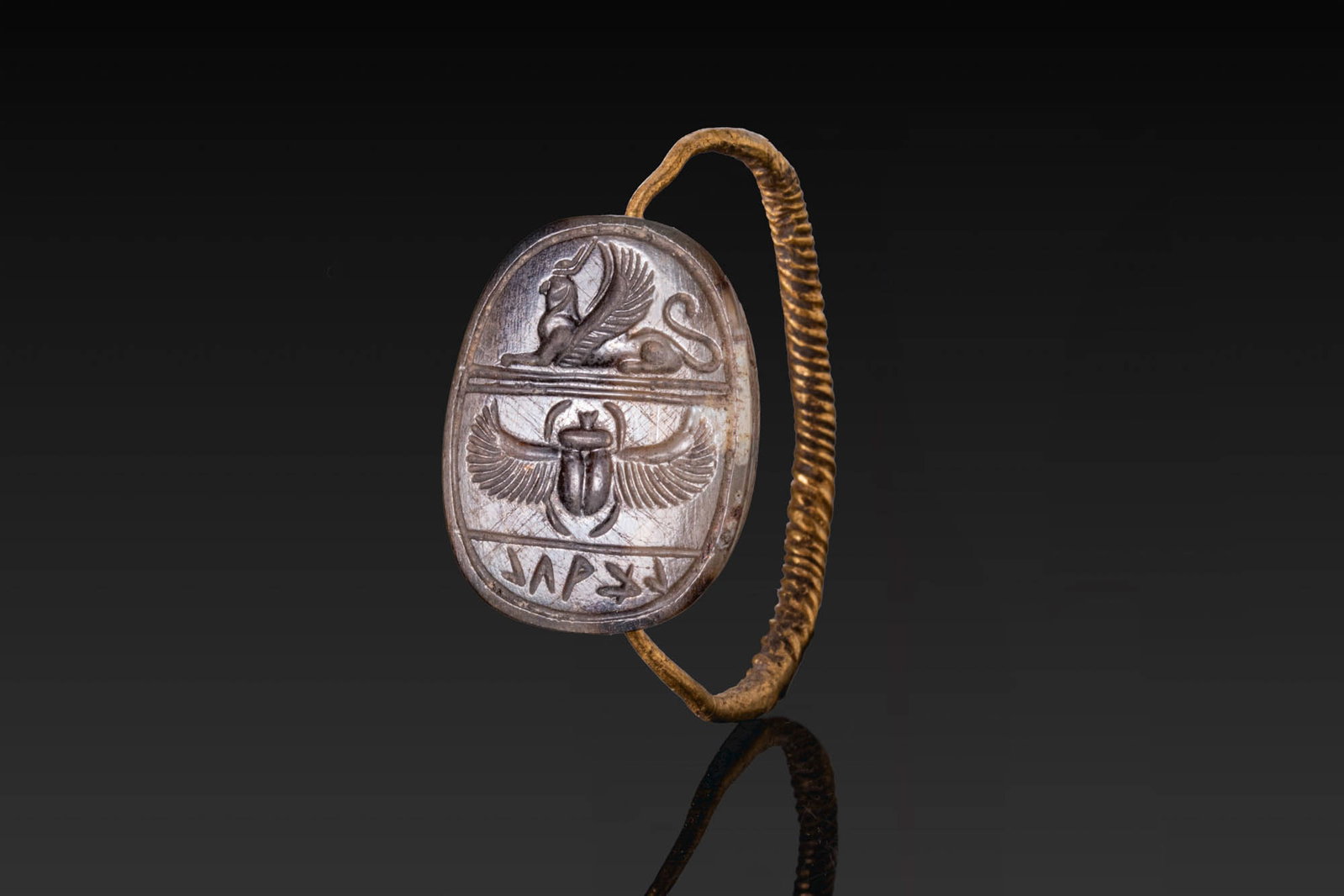 SYRO-PHOENICIAN GOLD SWIVEL RING WITH INSCRIBED STONE SCARAB: Ca. 664 - 30 BC. A gold swivel ring with a slender twisted hoop, set with a stone scarab carved with a rounded back, the head, clypeus, and wing cases in low relief, and a flat underside engraved with