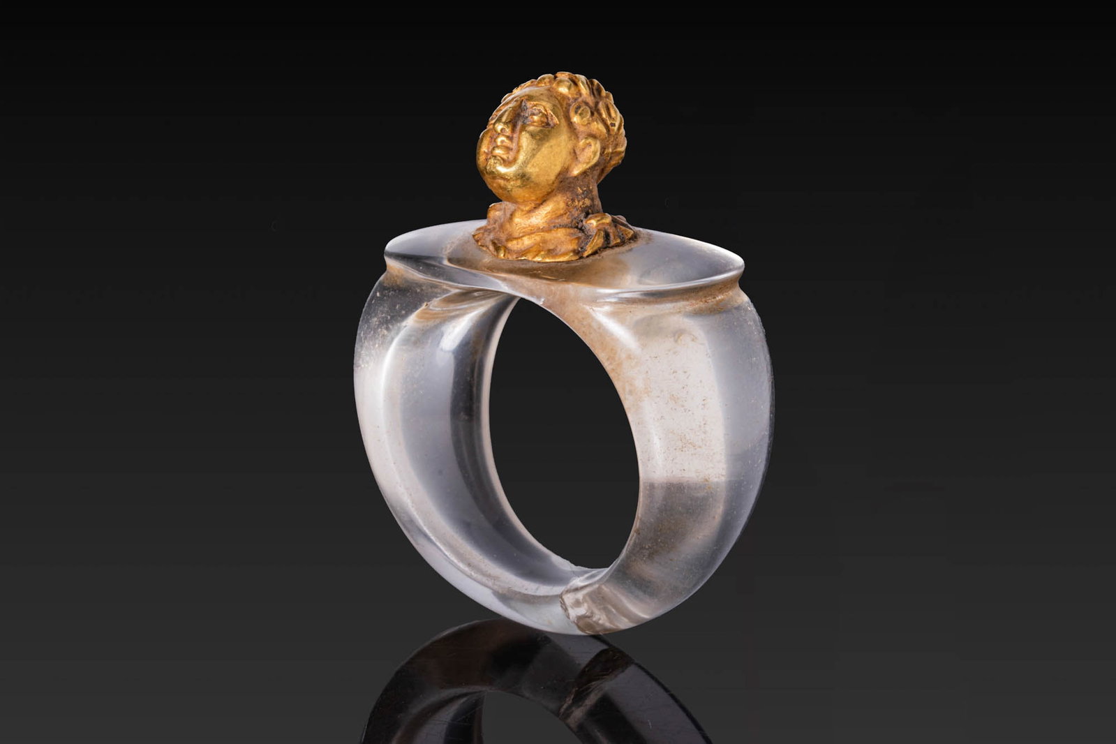 RARE ROMAN ROCK CRYSTAL AND GOLD RING WITH EMPEROR BUST: Ca. AD 1 - 300. A very rare rock crystal ring with a broad, rounded hoop and an oval bezel surmounted at the centre by a gold bust of an emperor, the head gently inclined, with short curling hair fram
