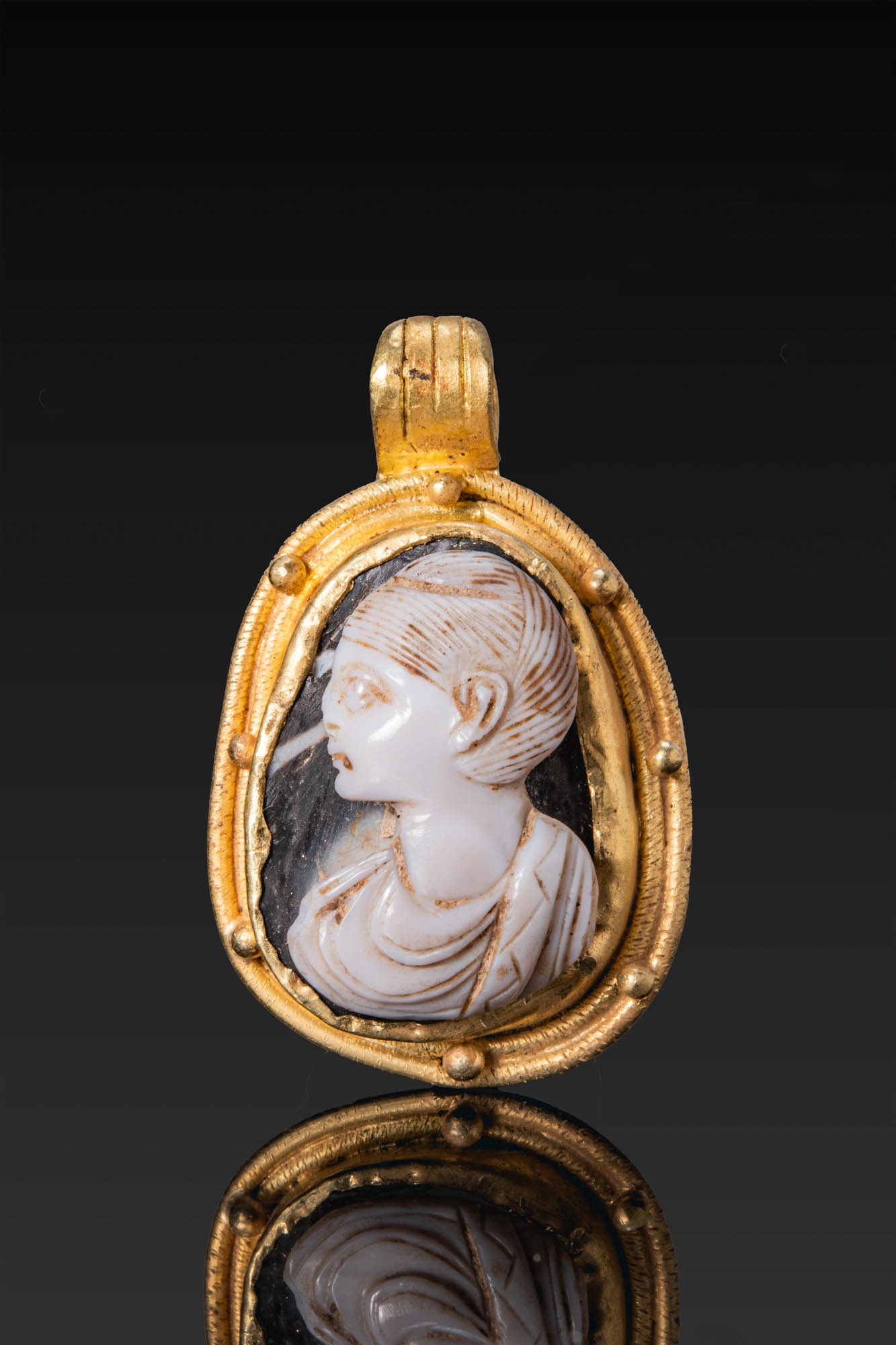 ROMAN AGATE CAMEO OF AN EMPRESS IN GOLD PENDANT: Ca. AD 1 - 300. A layered agate cameo carved with the bust of an empress in left profile, with combed hair and drapery over the shoulders, set in a gold pendant with an oval frame bordered by a row of