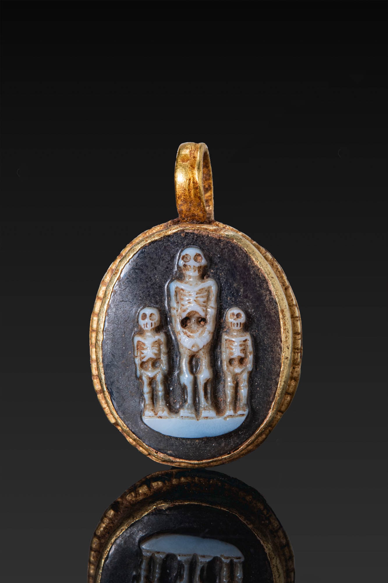 RARE ROMAN AGATE CAMEO OF MEMENTO MORI IN GOLD PENDANT: Ca. AD 1 - 300. A two-layered agate cameo carved in relief with three skeletons standing frontally on a groundline, the central figure larger and flanked by two smaller figures. The stone is set in an