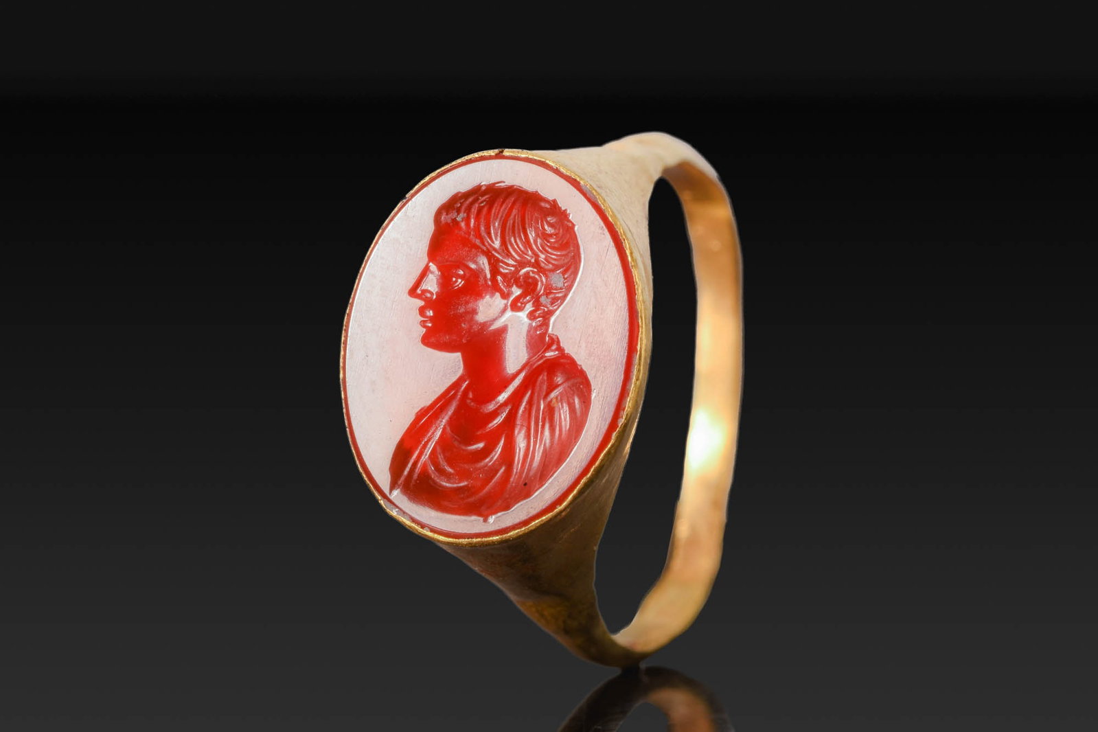 ROMAN GOLD RING WITH CARNELIAN INTAGLIO OF A YOUTH: Ca. AD 1 - 50. A gold ring with a slender hoop expanding to an oval bezel set with a carnelian intaglio engraved with a bust of a youth in profile to the left, with short wavy hair and drapery across 