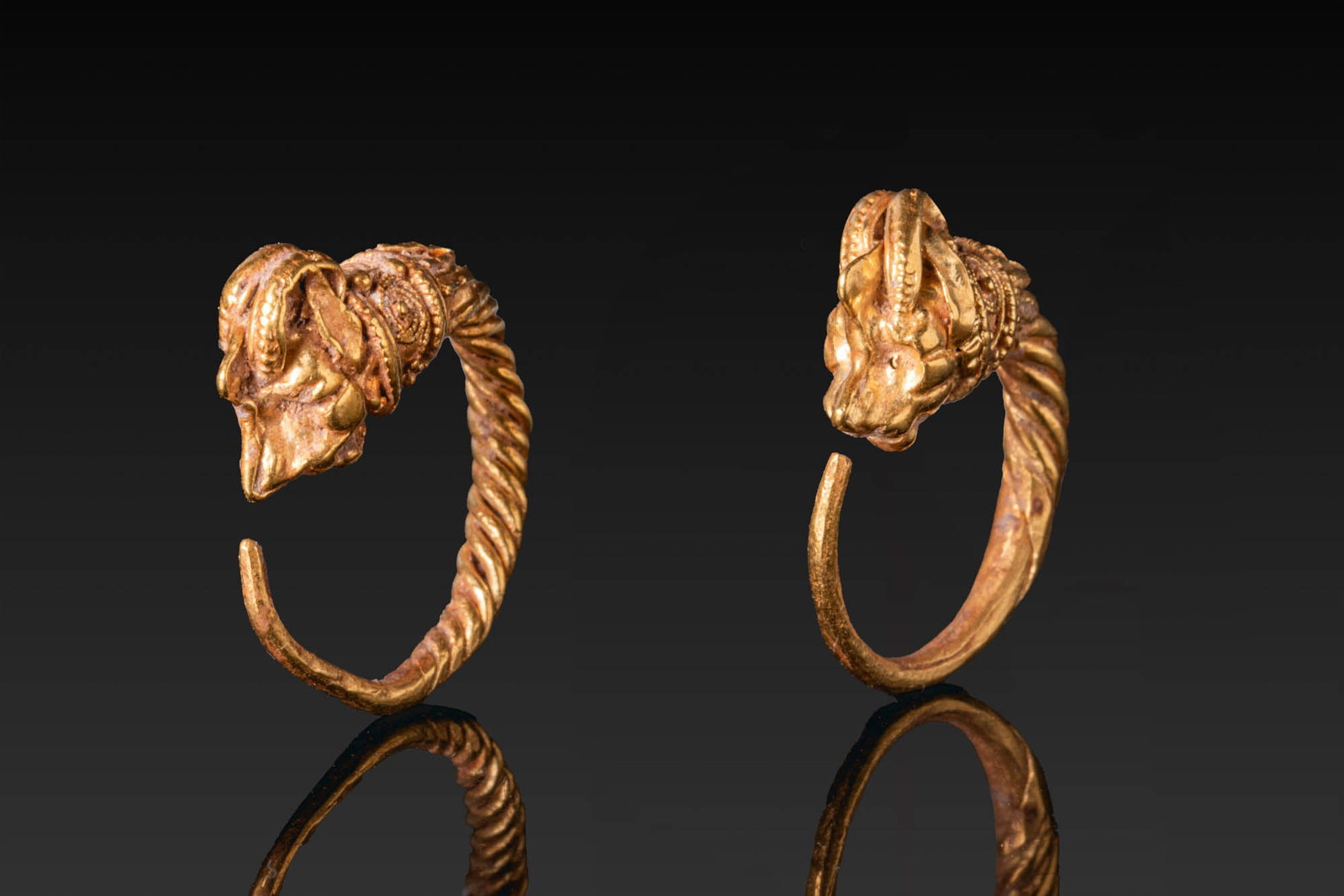 MATCHED PAIR OF HELLENISTIC GOLD IBEX-HEADED EARRINGS: Ca. 200 - 100 BC. A matched pair of gold earrings, each formed as a tapering twisted hoop with a hooked end at one side and an ibex head terminal at the other, set above a cylindrical collar decorated