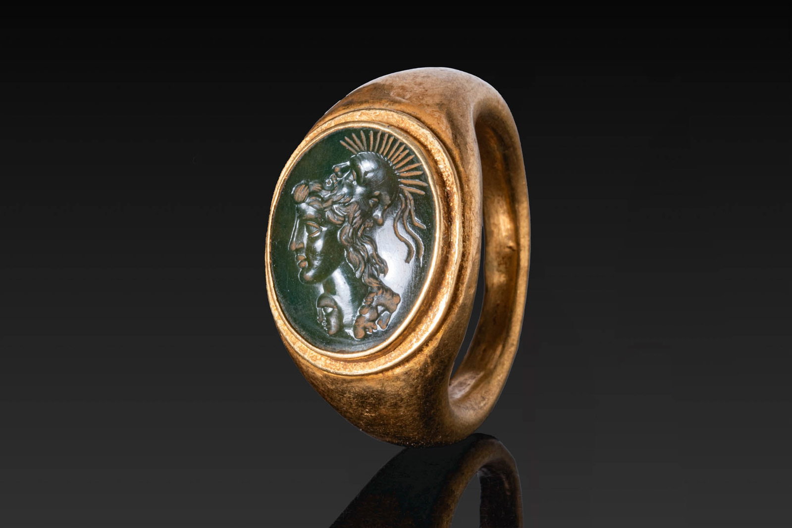 SUPERB ROMAN GREEN STONE INTAGLIO OF A GRYLLOS IN GOLD RING: Ca. AD 1 - 300. A dark green intaglio engraved with a gryllos composed of the profile head of a youth surmounted by a helmet with a radiate crest formed of two conjoined masks of bearded Silenus looki