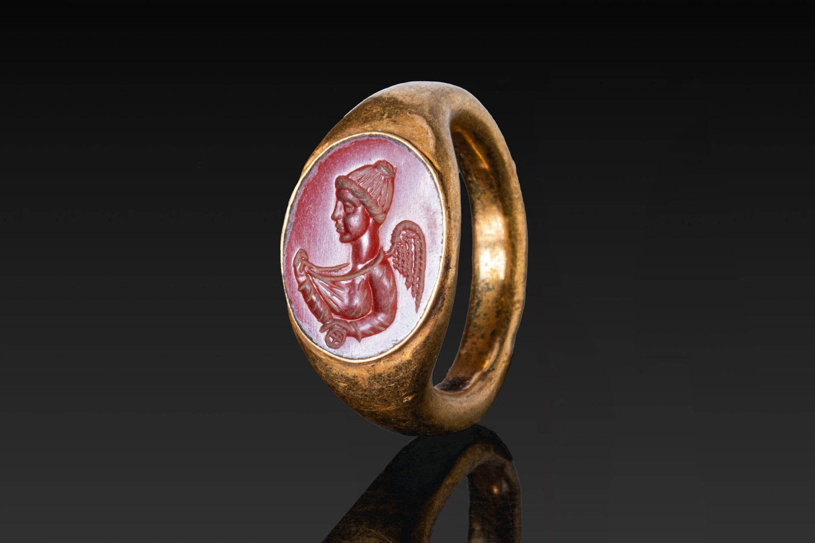 SUPERB ROMAN RED JASPER INTAGLIO OF FORTUNA IN GOLD RING: Ca. AD 1 - 300. A red jasper intaglio engraved with the left-facing bust of winged Fortuna, set in a possibly later gold ring with a rounded hoop and an oval bezel. For similar see: The Classical Art 
