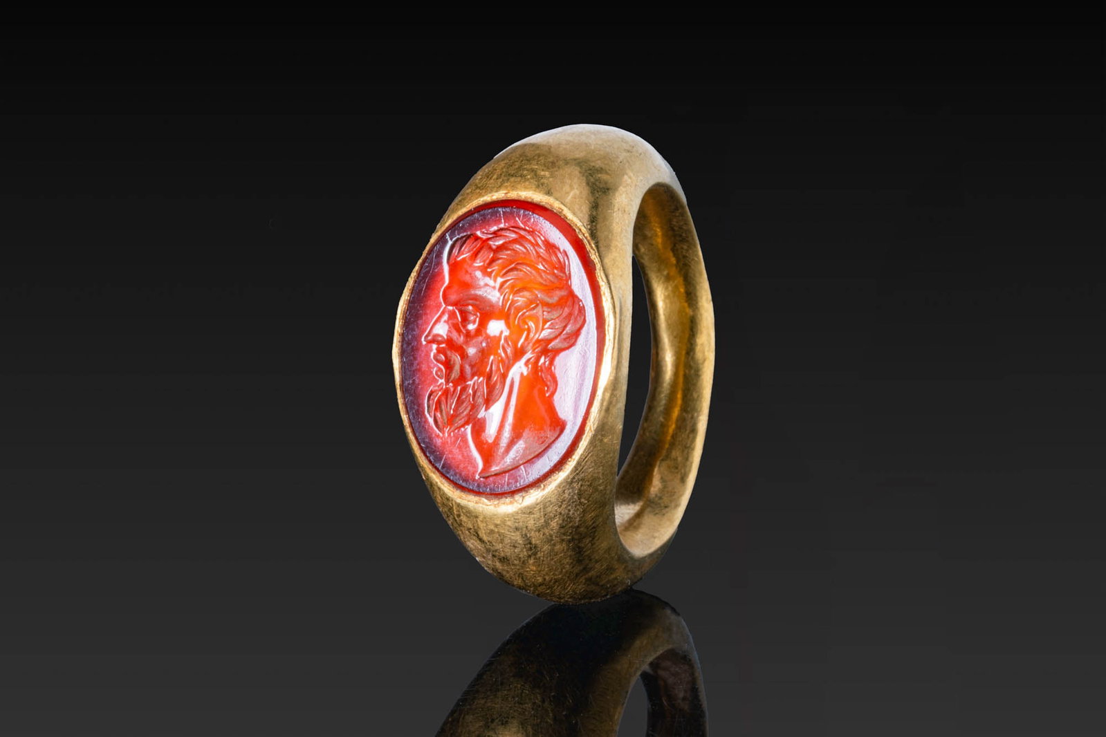 SUPERB ROMAN CARNELIAN INTAGLIO OF A PHILOSOPHER IN GOLD RING: Augustan Period, Ca. 33 BC - AD 14. A carnelian intaglio engraved with the profile head of a philosopher, with the hair brushed forward, a long beard, a pointed nose, and a muscular neck. It is set in