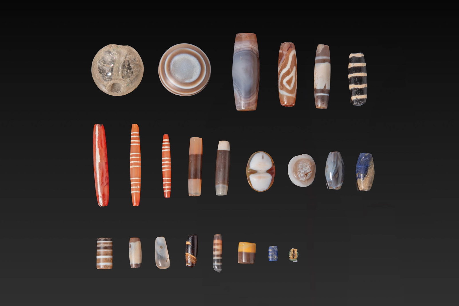 COLLECTION OF GREEK AND ROMAN STONE BEADS: Ca. 500 BC - AD 200. A collection of 23 stone beads, including banded agate, carnelian, and lapis lazuli examples in rounded, cylindrical, biconical and barrel shapes. Size: 8-52mm x 5-33mm; Weight: 1