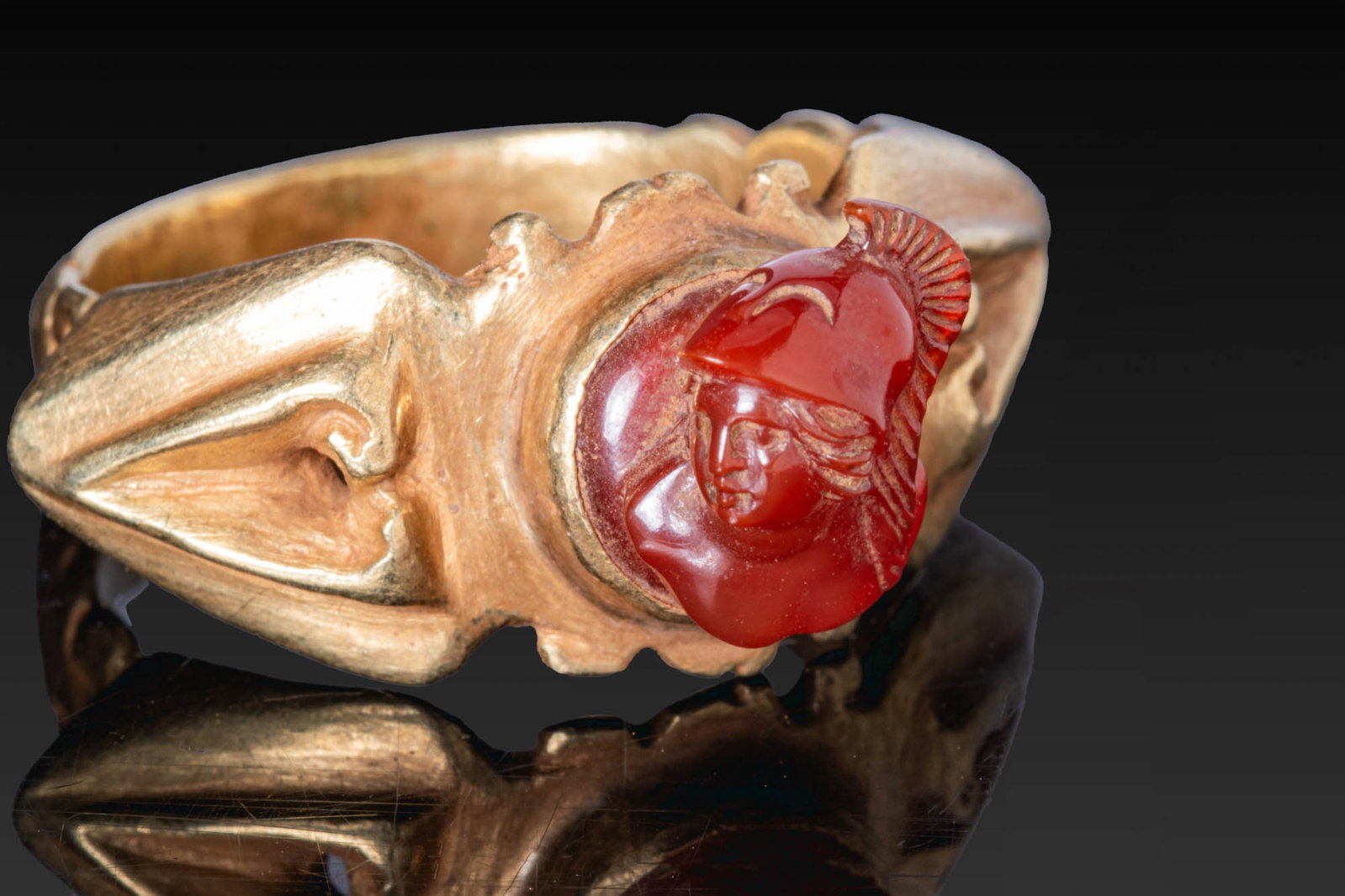 ROMAN CARNELIAN CAMEO BUST OF MINERVA IN GOLD RING: Ca. AD 100 - 200. A carnelian cameo carved with the head of Minerva in profile, wearing a crested helmet, set in a possibly later gold ring with a circular scalloped bezel, angular shoulders with rais