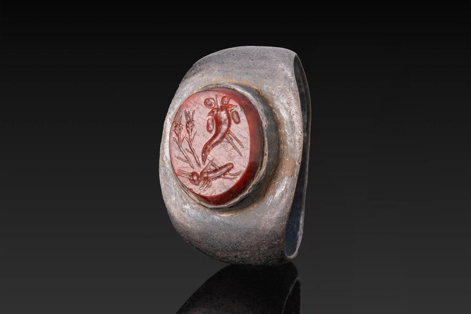 ROMAN RED JASPER INTAGLIO OF A CORNUCOPIA, WHEAT STALKS AND A GRASSHOPPER IN SILVER RING: Ca. AD 1 - 200. A red jasper intaglio engraved with a cornucopia beside two wheat stalks and a grasshopper below, set in a possibly later silver ring with a broad flat-section hoop widening towards