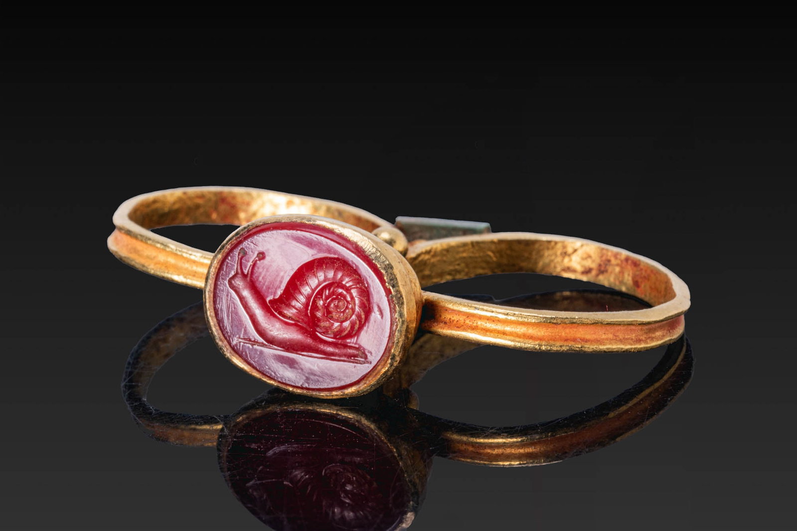 ROMAN GOLD DOUBLE RING WITH CARNELIAN INTAGLIO OF A SNAIL: Ca. AD 1 - 300. A gold double ring formed of two hoops joined beneath an oval bezel cell with a carnelian intaglio engraved with a snail on a groundline, with small gold granules at the junction and