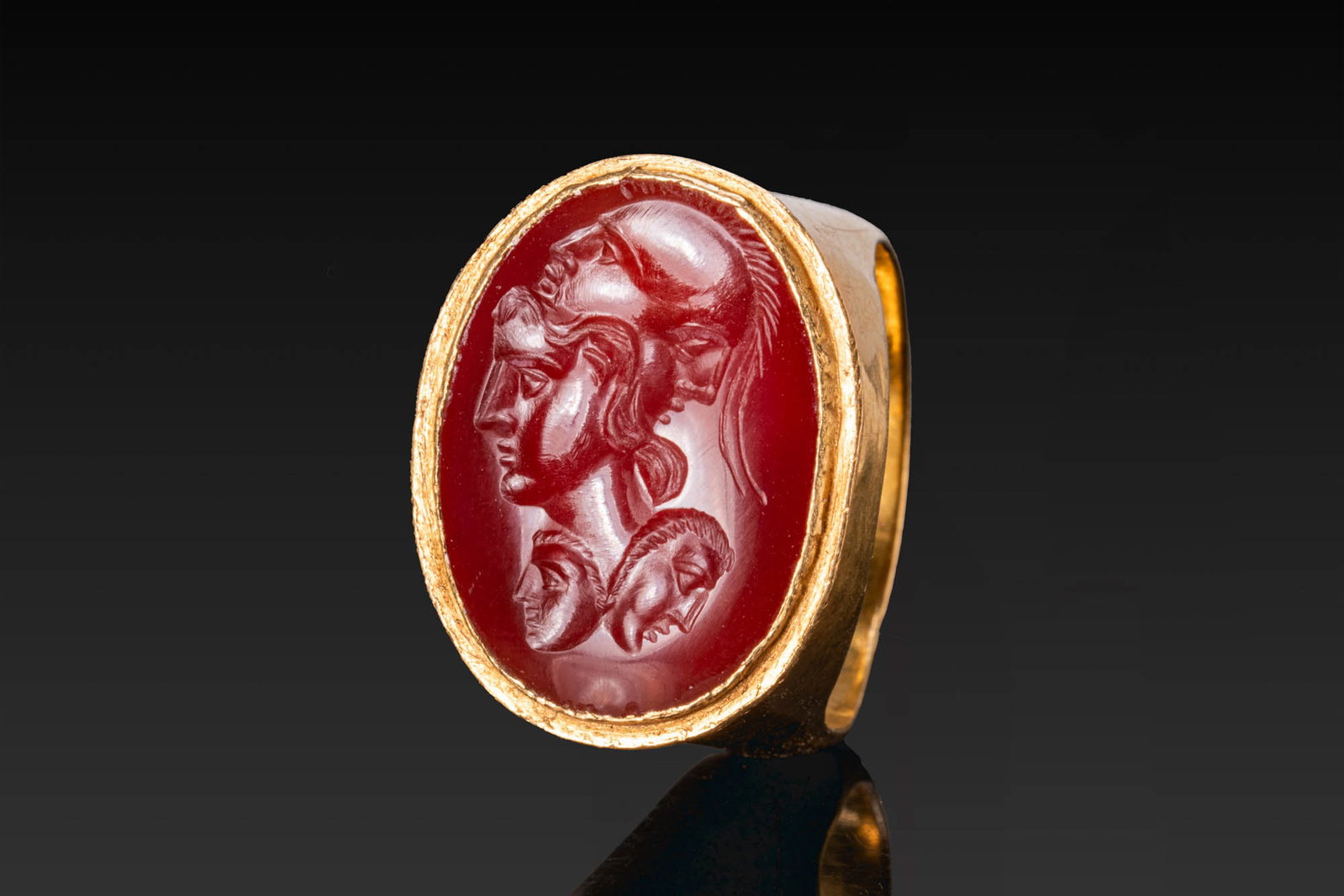 ROMAN CARNELIAN INTAGLIO OF MINERVA AND JANIFORM HELMET IN GOLD RING: Ca. AD 300. A carnelian intaglio engraved with the head of Minerva in left profile, wearing a janiform helmet with a crested crown, and with two smaller masks below. It is mounted in a possibly later 