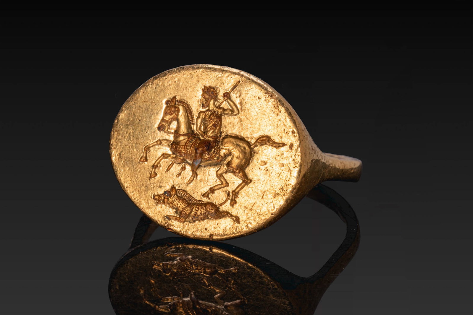 SUPERB ROMAN SOLID GOLD RING OF A GOD HUNTING A WILD BOAR: Ca. AD 1 - 100. A gold ring with a slender hoop expanding sharply at the shoulders to a broad oval bezel engraved with a mounted god riding to the left, his horse in full stride and the god holding a