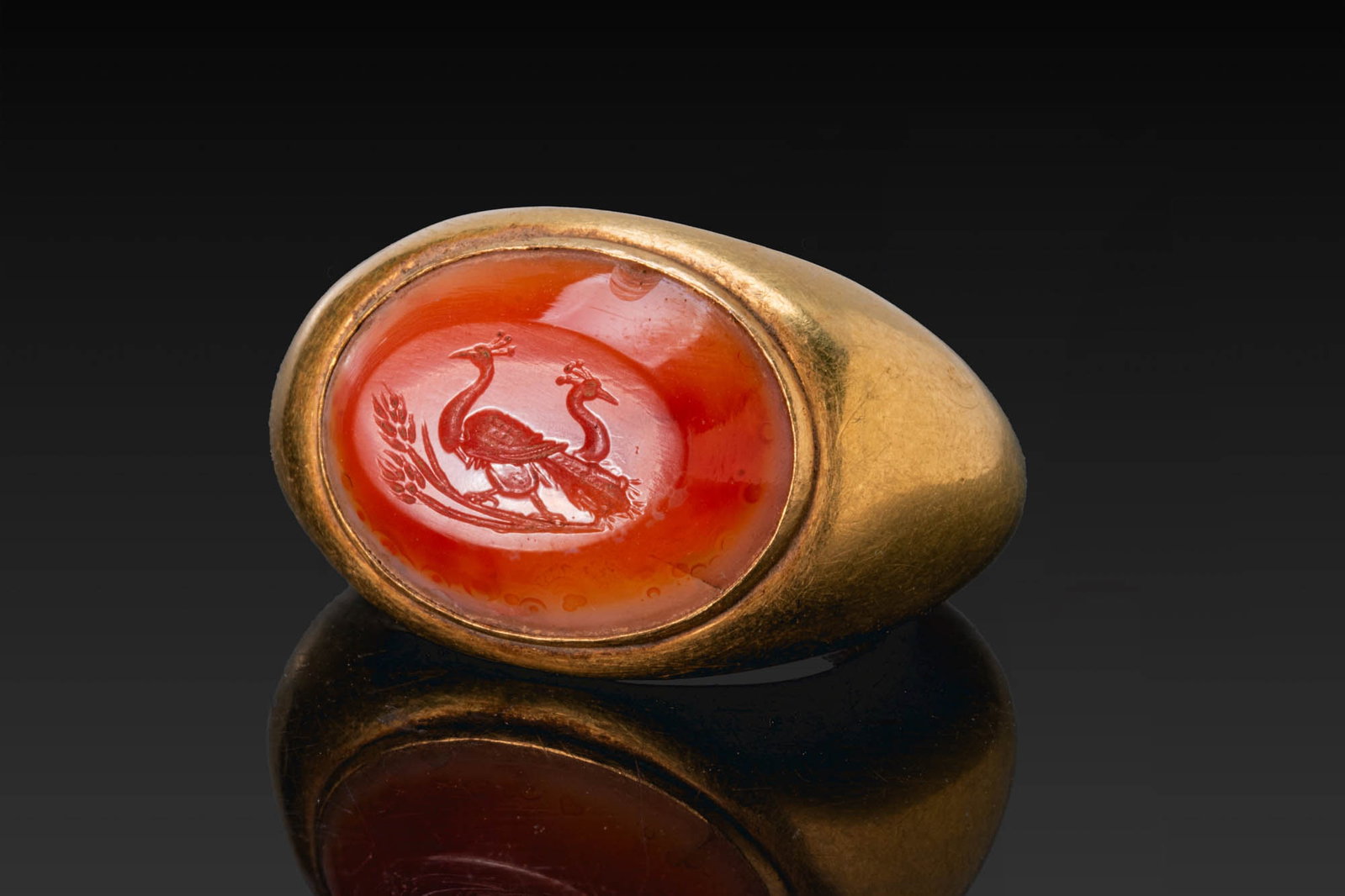 LARGE ROMAN GOLD RING WITH AGATE INTAGLIO OF PEACOCKS ON WHEAT STALKS: Ca. AD 1 - 50. A gold ring with a broad rounded hoop expanding to a wide oval bezel set with a convex agate intaglio engraved with a pair of peacocks standing above wheat stalks. For similar see: The