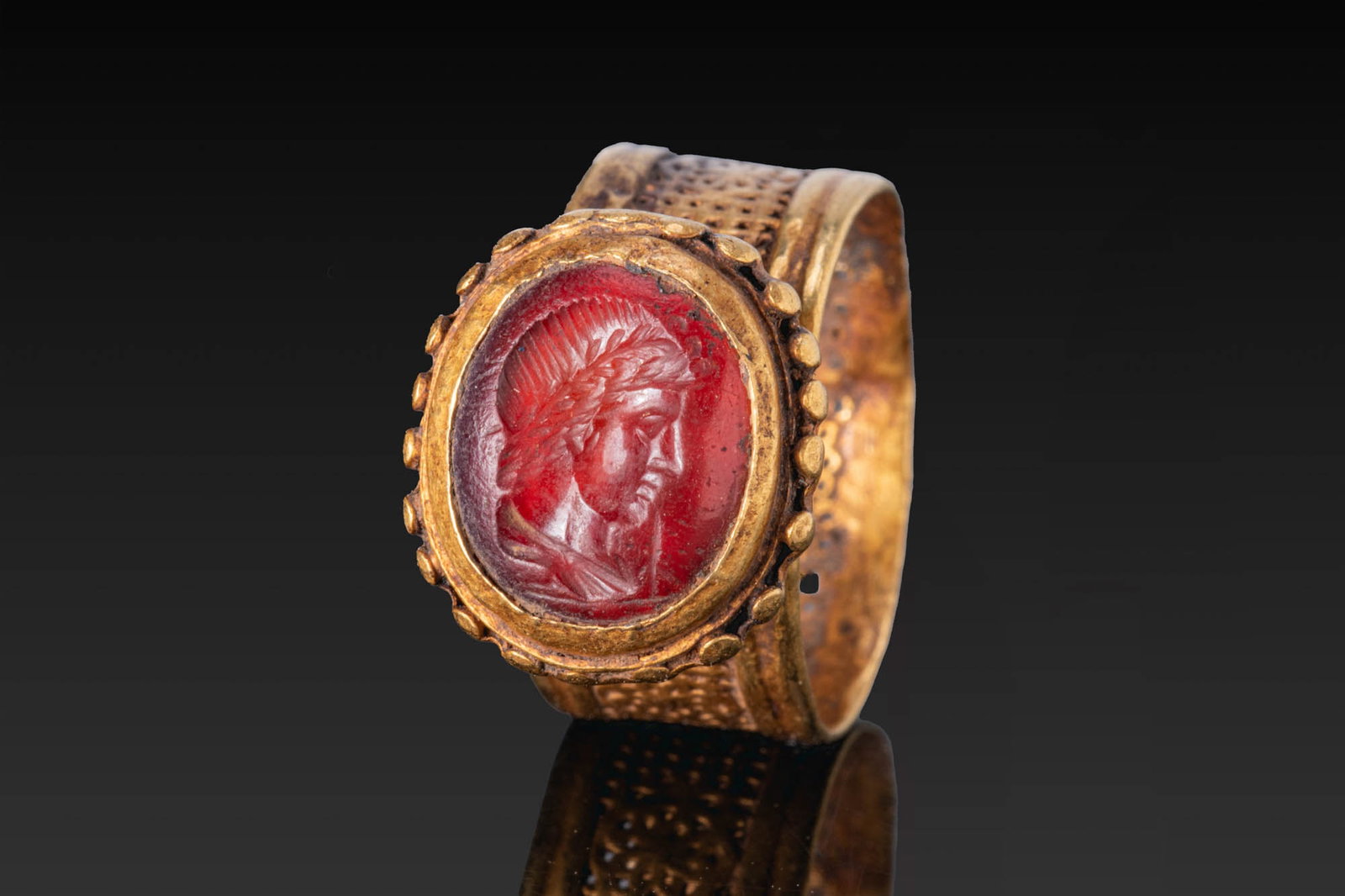 LATE ROMAN GOLD OPENWORK RING WITH CARNELIAN INTAGLIO OF APOLLO: Ca. AD 400 - 500. A gold ring with a broad openwork hoop pierced with rows of small circular holes and decorated with a rosette motif. The high bezel has a fluted underside and a border of bosses fram