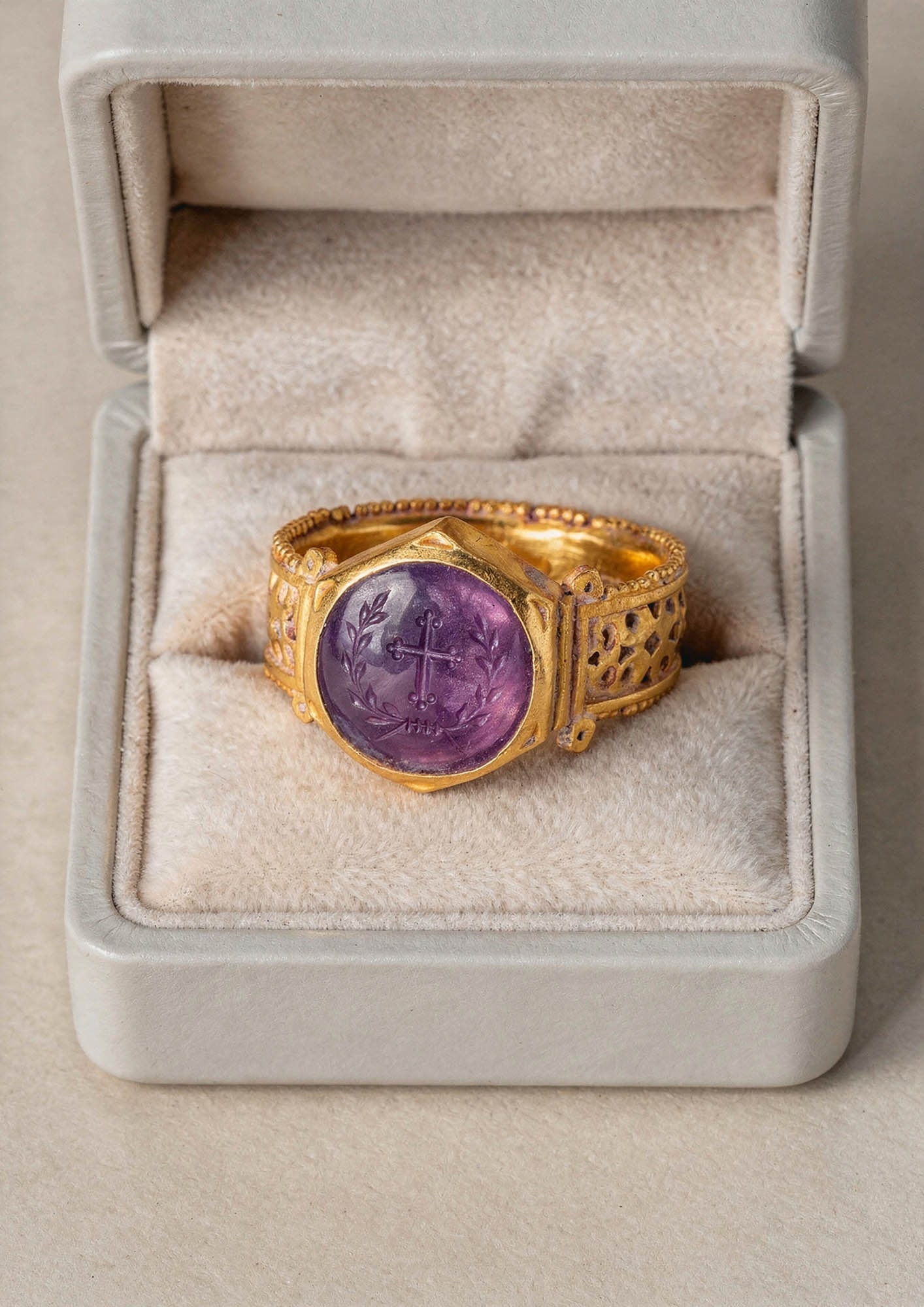 LATE ROMAN GOLD OPENWORK RING WITH AMETHYST INTAGLIO OF A CROSS IN A LAUREL WREATH: Ca. AD 300 - 400. A gold ring with a broad openwork hoop pierced with crosses formed of triangular arms with lozenge centres, with circles in the quadrants, all between beaded borders, and a hexagonal