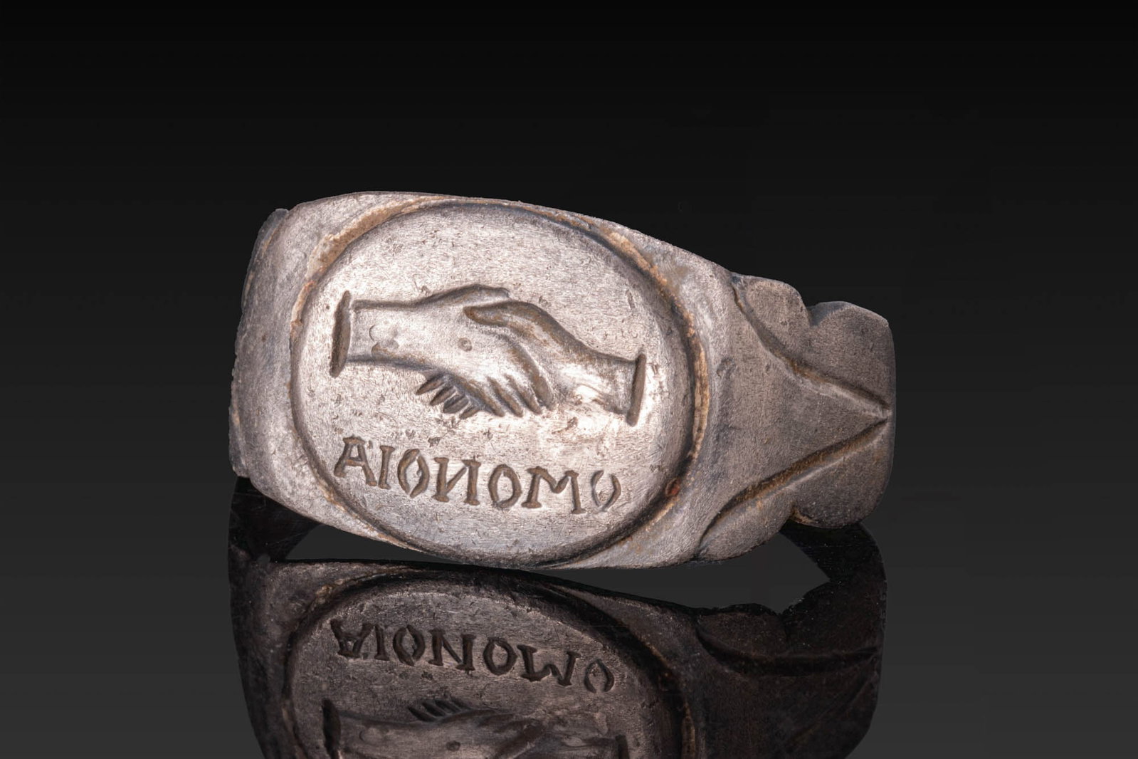 ROMAN SILVER RING WITH CLASPED HANDS AND OMONOIA INSCRIPTION: Ca. AD 1 - 300. A silver ring with a flat-section hoop, lobed shoulders incised with V-shaped motifs, and a large oval bezel engraved with clasped right hands above the inscription OMONOIA, meaning "h