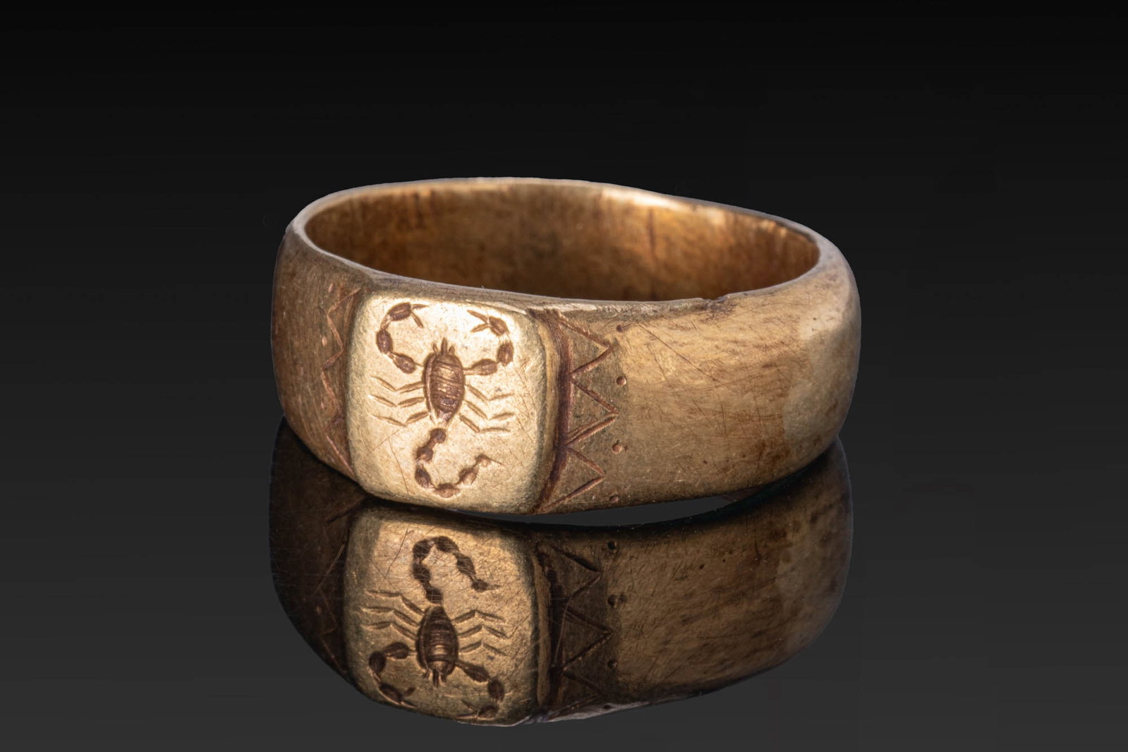 ROMAN GOLD RING WITH SCORPION: Ca. AD 1 - 200. A gold ring with a broad D-section hoop widening to a slightly raised rectangular bezel engraved with a scorpion. Zigzag bands and rows of punched dots frame the bezel. For similar