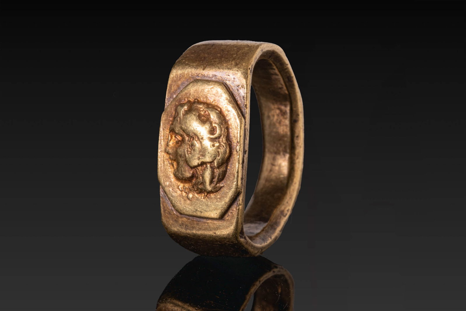 ROMAN GOLD RING WITH ALEXANDER THE GREAT AS HERAKLES: Ca. AD 1 - 200. A gold ring with a broad flat-section polygonal hoop and a flat rectangular bezel with an octagonal plate in low relief with the head of Alexander the Great depicted as Herakles in lef