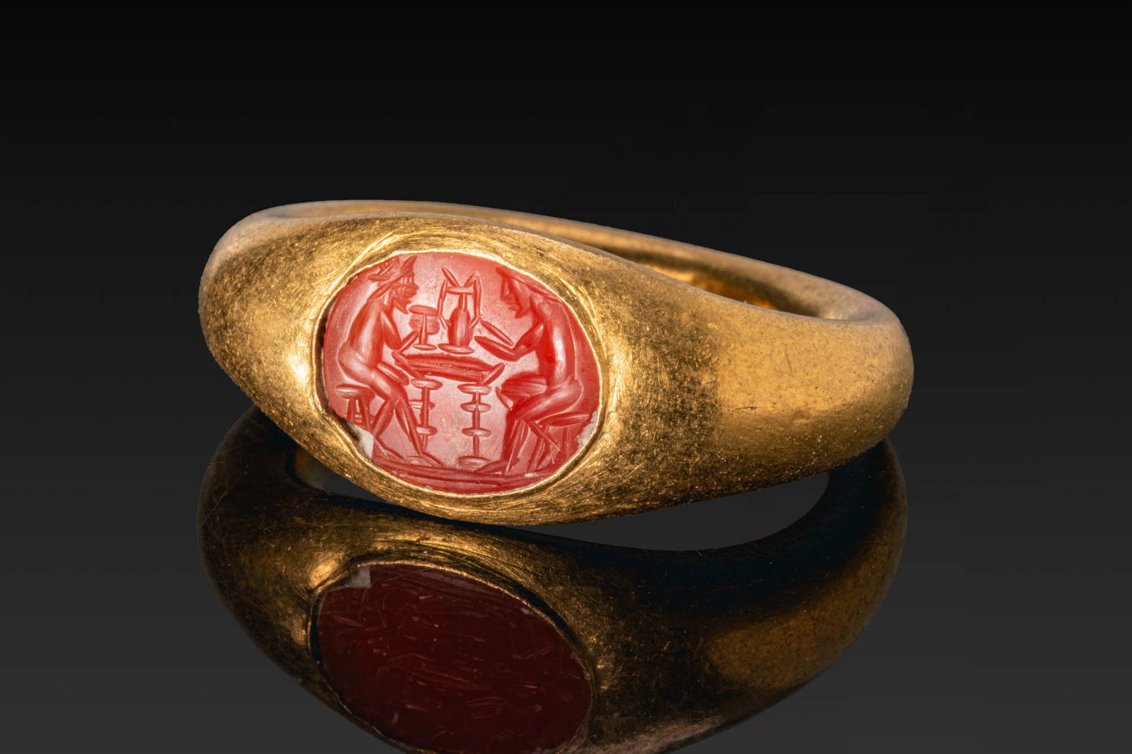 ROMAN GOLD RING WITH CARNELIAN INTAGLIO OF A DIONYSIAC SCENE: Ca. AD 50 - 200. A gold ring with a plain, rounded hoop and an oval bezel set with a carnelian intaglio engraved with two seated figures facing one another across a table, each holding a vessel. For