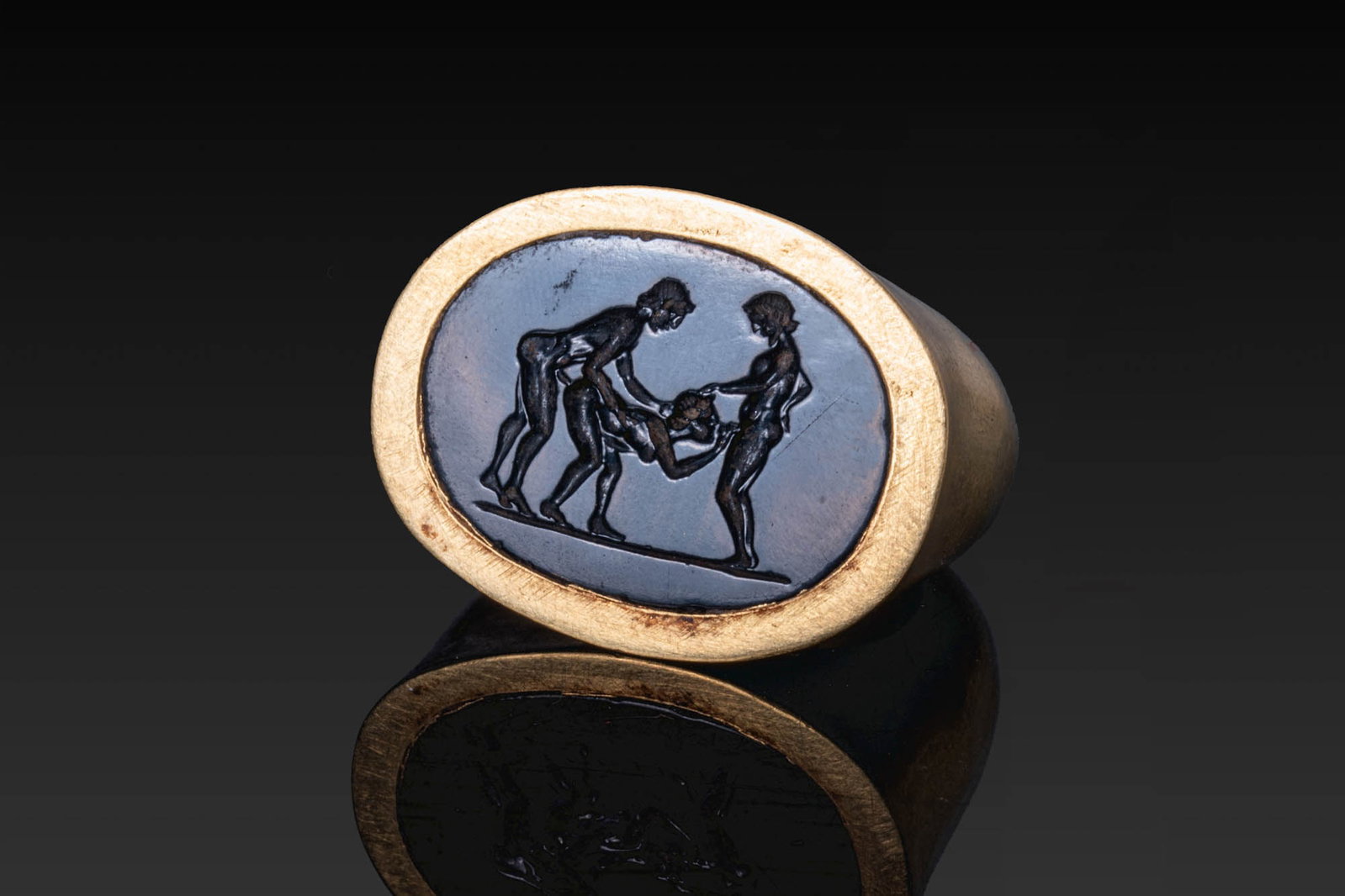 VERY RARE ROMAN GOLD RING WITH BLACK STONE INTAGLIO OF AN EROTIC SCENE: Ca. 100 BC - AD 100. A black stone intaglio engraved with an erotic scene showing a nude woman bent between two nude men, on a groundline, set in a possibly later gold ring with a broad, rounded hoop 