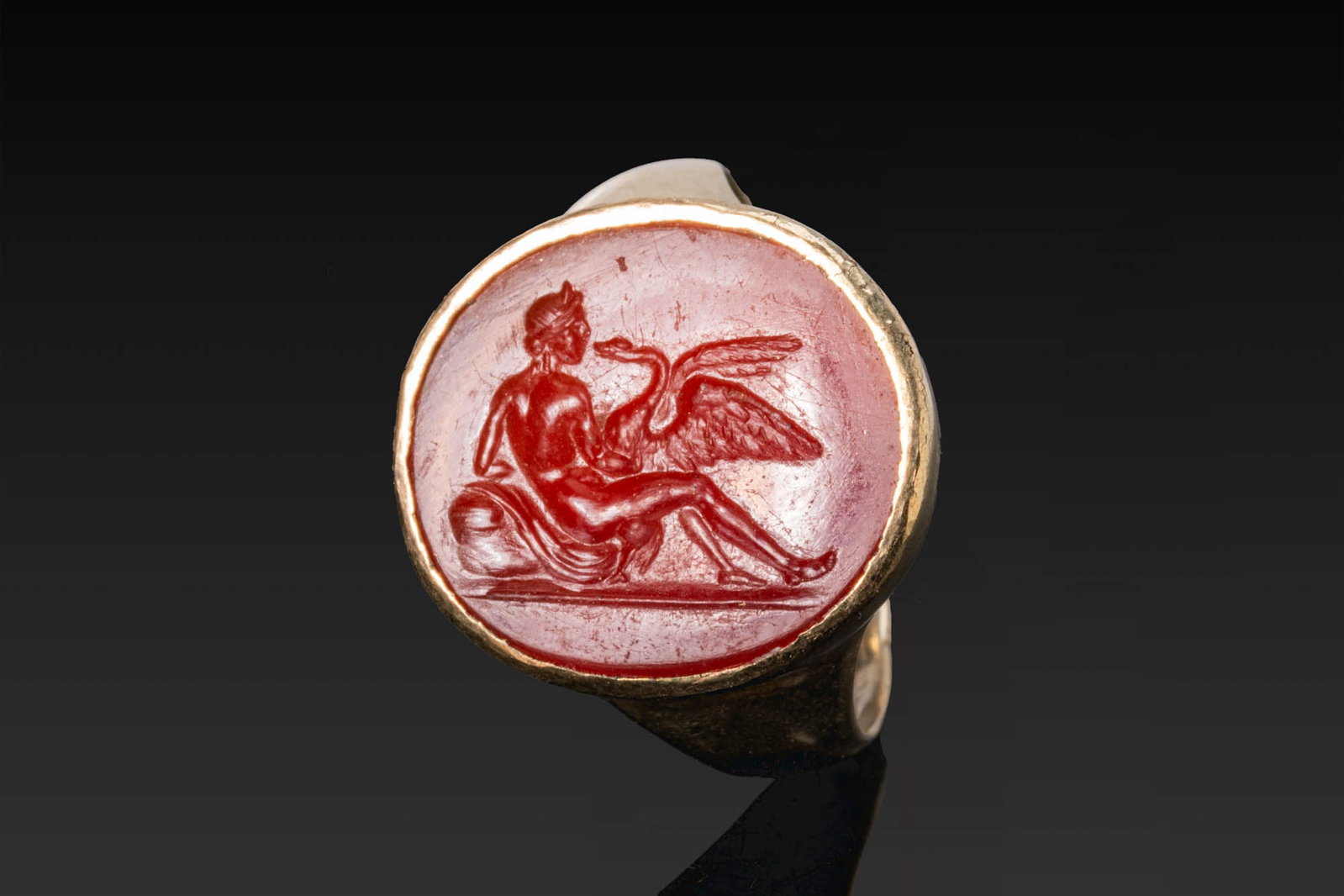 RARE ROMAN CARNELIAN INTAGLIO OF LEDA AND SWAN IN GOLD RING: Ca. AD 1 - 300. A carnelian intaglio engraved with reclining Leda beside the swan, set in a possibly later gold ring with a circular bezel and a plain hoop tapering from the shoulders. For similar see