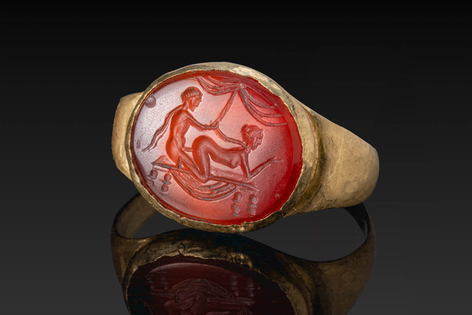 ROMAN GOLD RING WITH CARNELIAN INTAGLIO OF AN EROTIC SCENE: Ca. AD 100 - 300 (or later). A gold ring with a tapering hoop and an oval bezel set with a carnelian intaglio engraved with an erotic scene of a couple beneath drapery, the female figure on all fours 