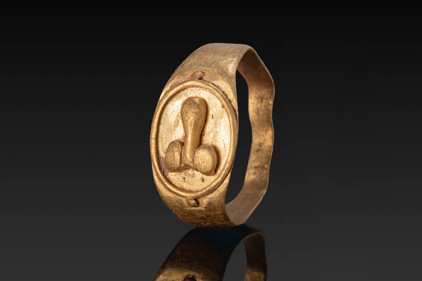 LATE ROMAN GOLD RING WITH PHALLUS: Ca. AD 200 - 400. A gold ring with a flat-section tapering hoop and an oval bezel plate enclosed by a raised border with pellets above and below, centred on an applied phallus. For similar see: The Br