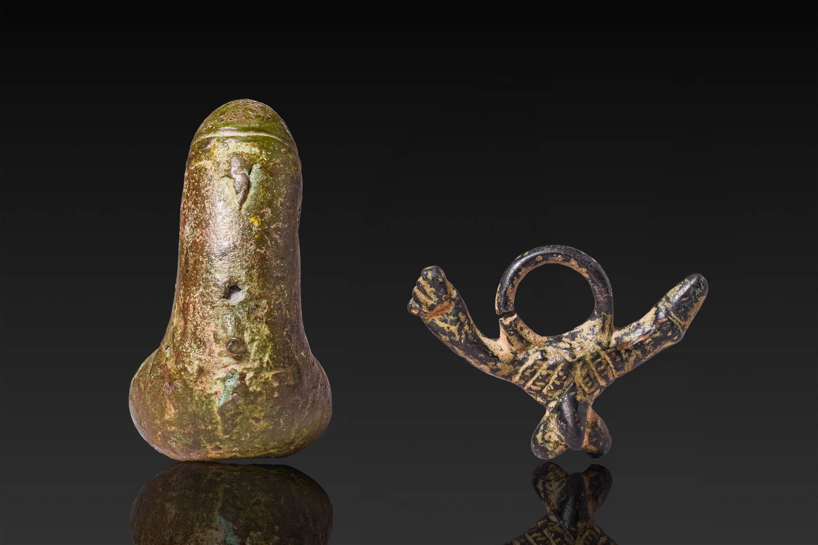 PAIR OF ROMAN BRONZE PHALLIC AMULETS: Ca. AD 1 - 300. A pair of bronze phallic amulets, including, from left to right: a phallus with a rounded glans, a cylindrical shaft, testicles, and perforations through the body; and a phallic pendan