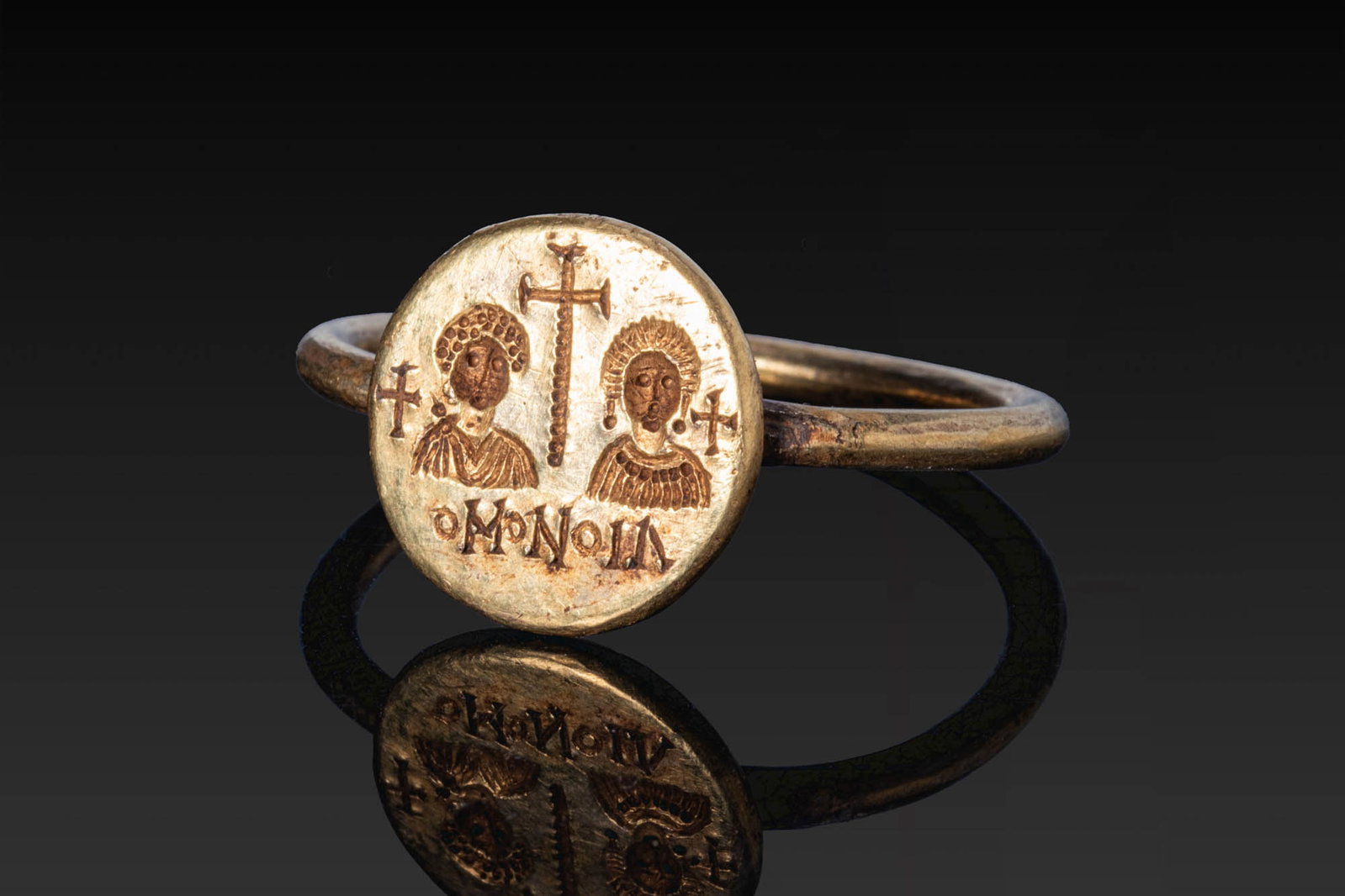 RARE BYZANTINE GOLD MARRIAGE RING: Ca. AD 600. A gold ring with a slender, round-section hoop and a circular bezel engraved with the frontal busts of a bride and groom beneath a tall cross, flanked by smaller crosses, with the inscript