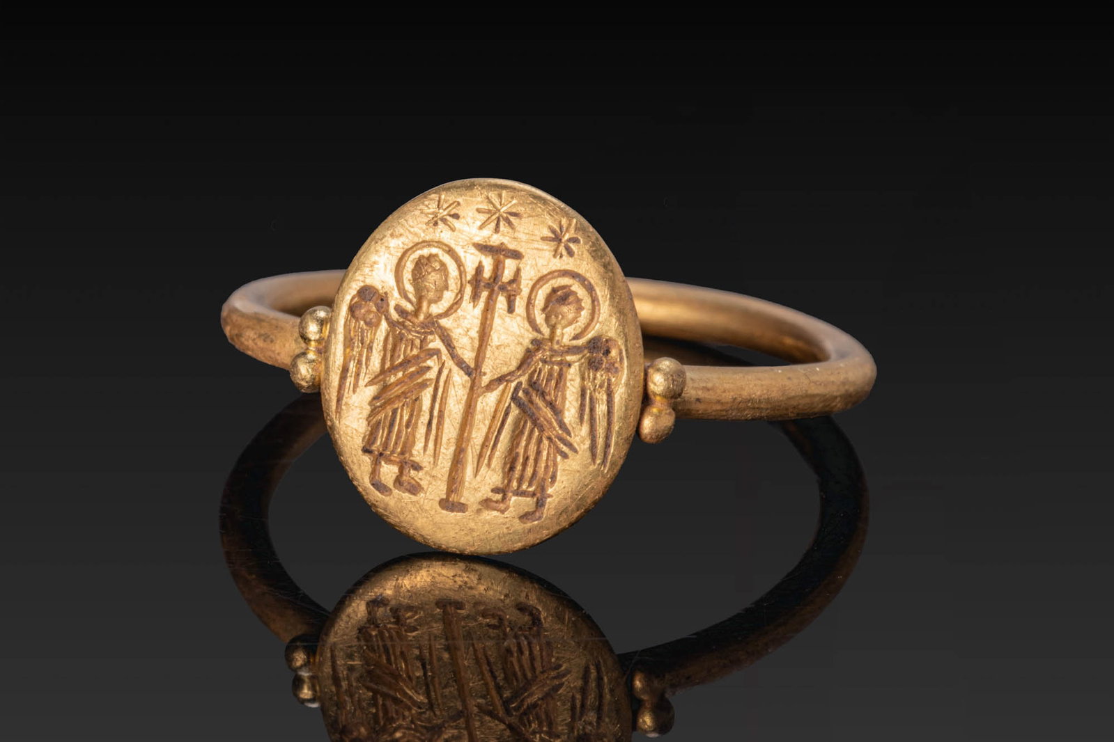 BYZANTINE GOLD RING WITH CENTRAL CROSS BETWEEN ARCHANGELS AND STARS: Ca. AD 600. A gold ring with a plain round-section hoop and an oval bezel, flanked at each side by a pair of granules. The bezel is engraved with two haloed archangels standing in profile on either si