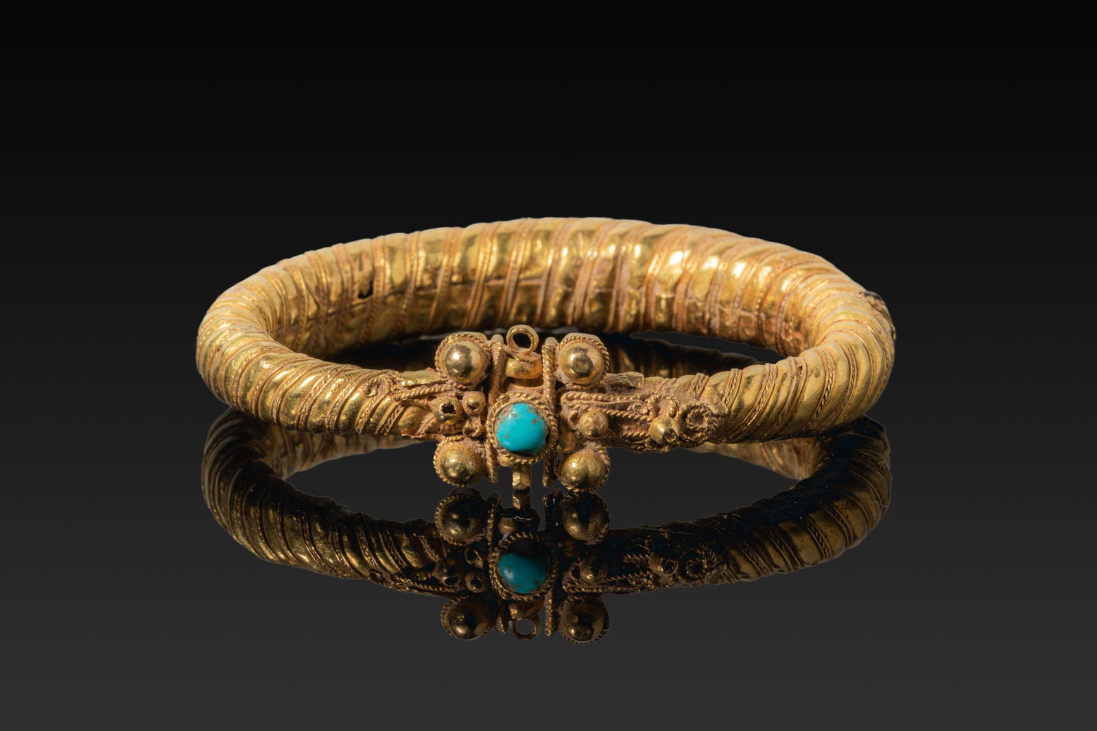 BYZANTINE GOLD ARMLET WITH CENTRAL TURQUOISE: Ca. AD 1000. An armlet made from sheet gold folded into a hollow tapering hoop with alternating ribs and ropework bands. The front section is decorated with scrollwork and filigree panels leading to a