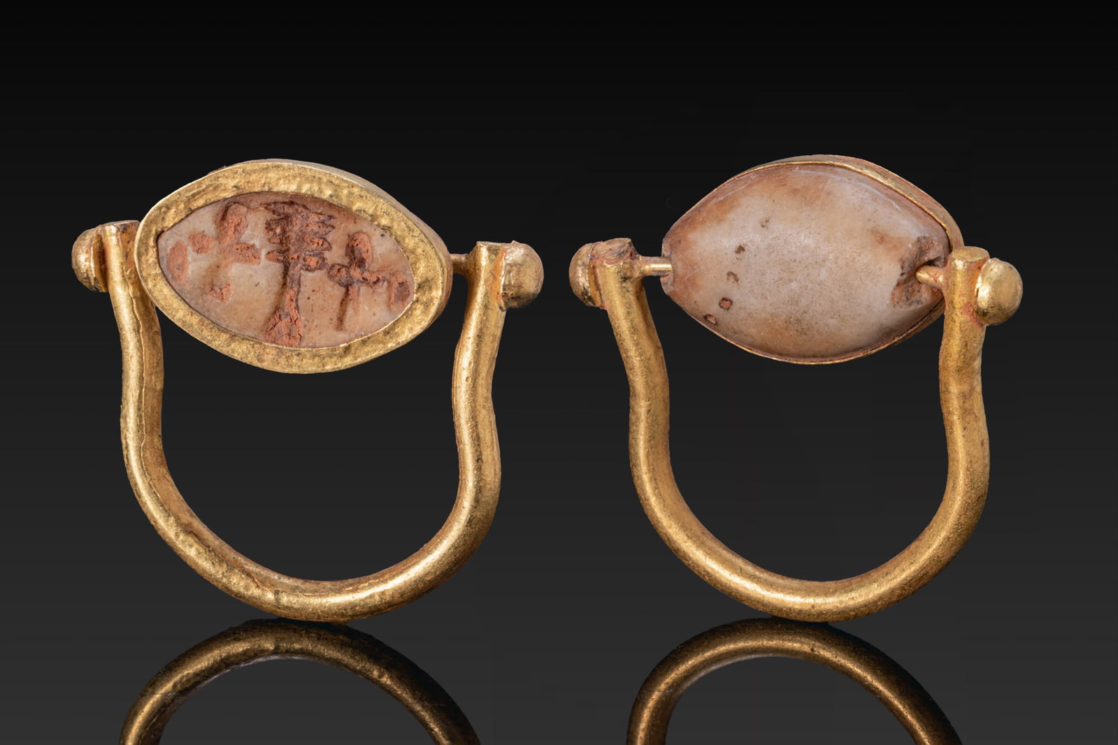 EGYPTIAN STONE COWROID WITH DJED PILLAR BETWEEN ANKHS IN GOLD SWIVEL RING: New Kingdom, Ca. 1550 - 1069 BC. A stone cowroid with a smooth convex upper face and a flat underside engraved with a djed pillar between ankh signs. Set in a possibly later gold swivel ring with a ro