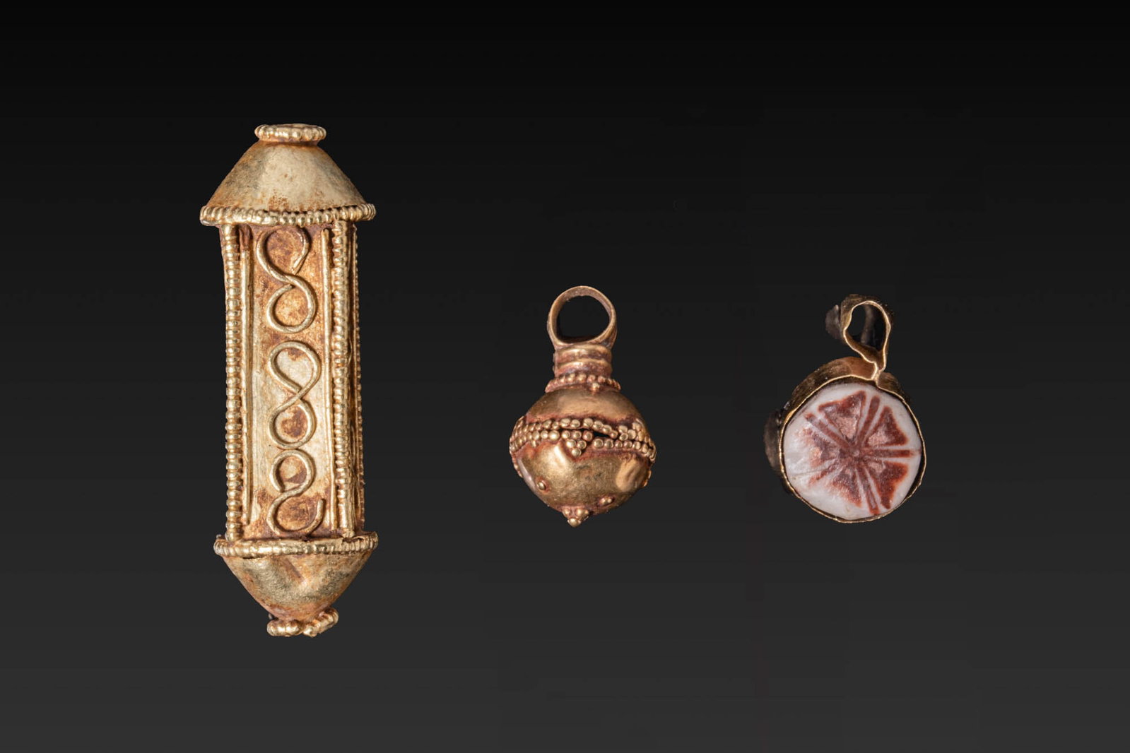 COLLECTION OF GREEK GOLD AND GLASS PENDANTS: Ca. 800 - 600 BC. A collection of three pendants, including, from left to right: a long pendant with tapering conical terminals and side panels framed by beaded borders, each decorated with a sinuous 