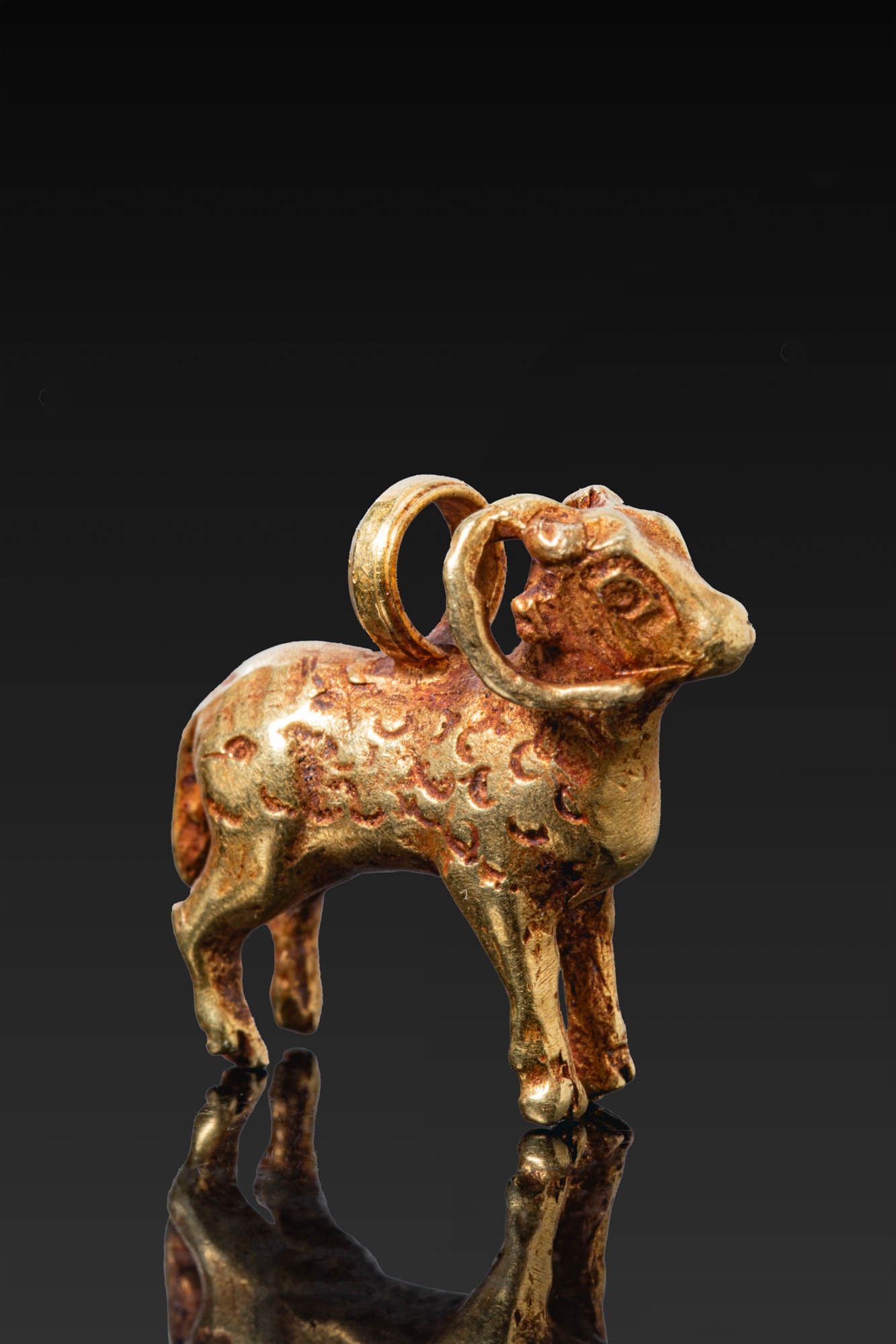 GREEK GOLD RAM-SHAPED PENDANT: Ca. 500 BC. A gold pendant in the form of a ram standing on four slender legs, with curved horns, a short tail, and a body with punched spiral motifs imitating fleece. A large suspension loop is set o