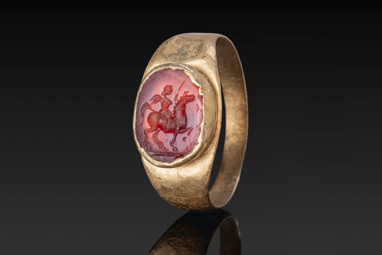 GREEK CARNELIAN INTAGLIO OF EROS RIDING A HORSE IN GOLD RING: Ca. 400 BC. A carnelian intaglio engraved with winged Eros riding a horse to the right, holding a whip, mounted in a possibly later gold ring with a flat-section tapering hoop and an oval bezel. For s