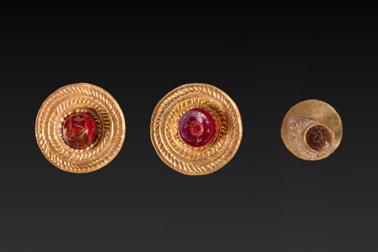 PAIR OF GREEK GOLD AND GARNET APPLIQUES: Ca. 300 - 100 BC. A pair of gold appliques, each with a circular upper section decorated with concentric herringbone bands around a central garnet boss, fitted on the reverse with a tubular attachment